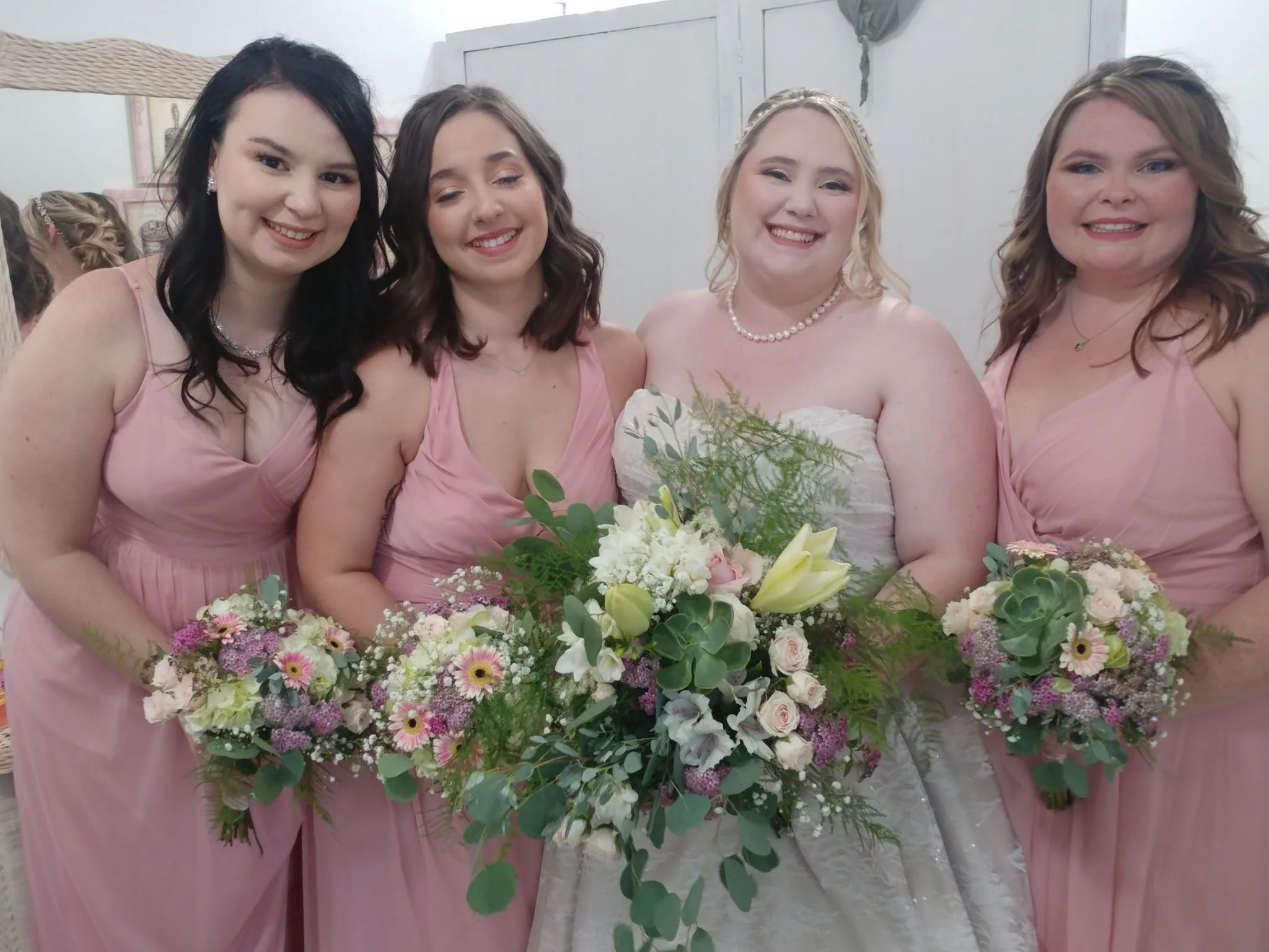 A bride in a white wedding dress holding a bouquet of white and pink flowers, standing with four bridesmaids in pink dresses, each holding smaller bouquets of pink and white flowers, all smiling.