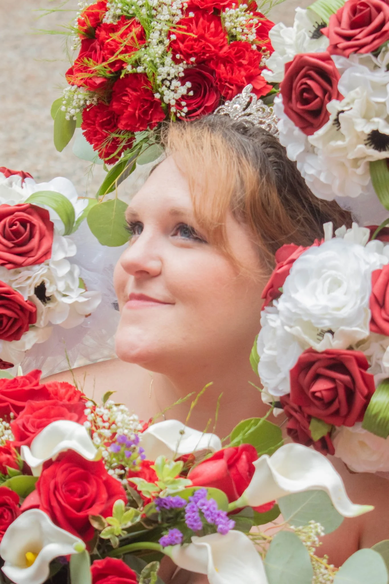 A woman with red hair surrounded by a large arrangement of red, white, and purple flowers, looking upwards with a gentle smile.