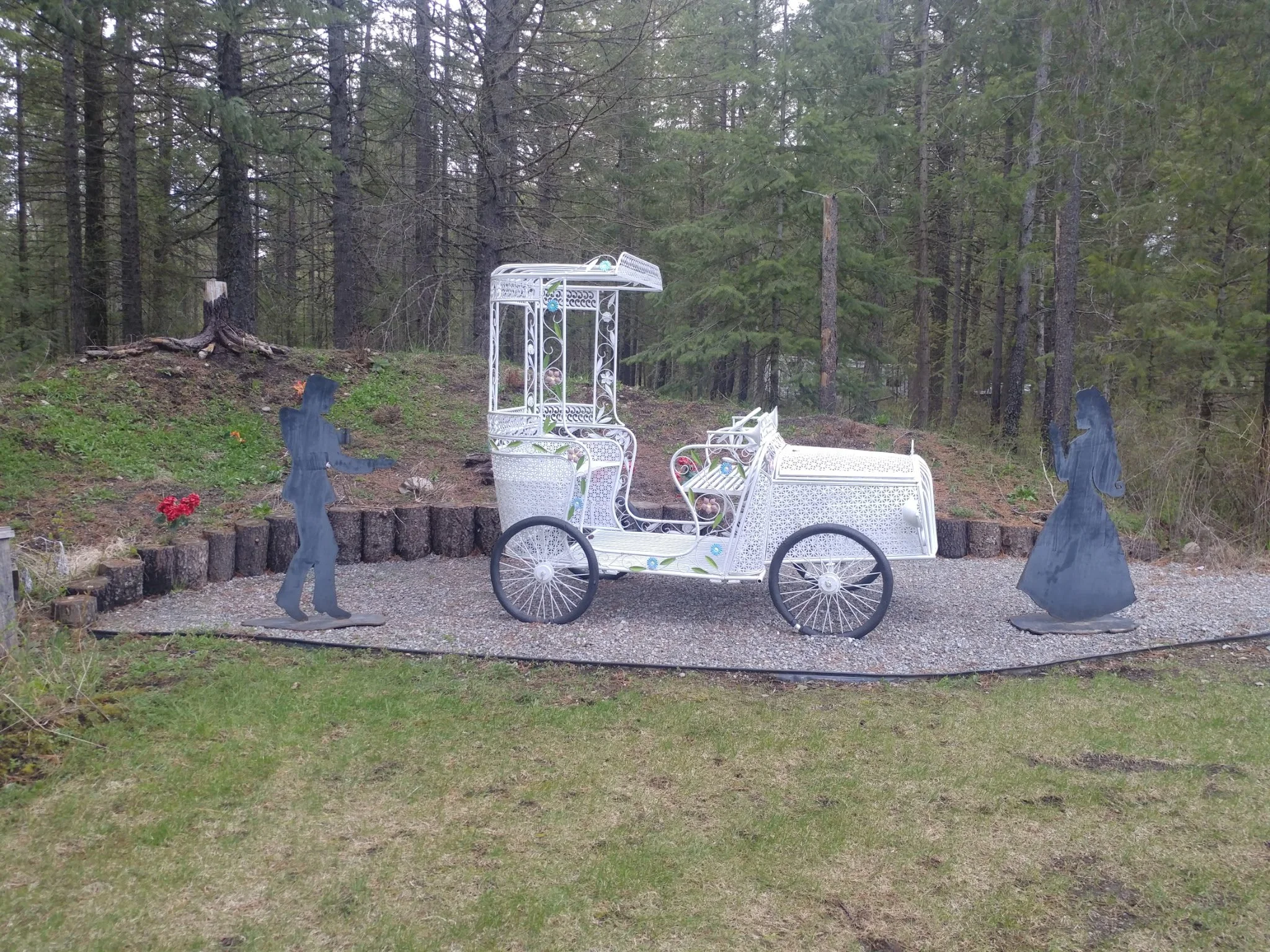 Decorative white carriage with ornate metalwork, surrounded by cutouts of a man and woman silhouette, set in a garden area with trees in the background.