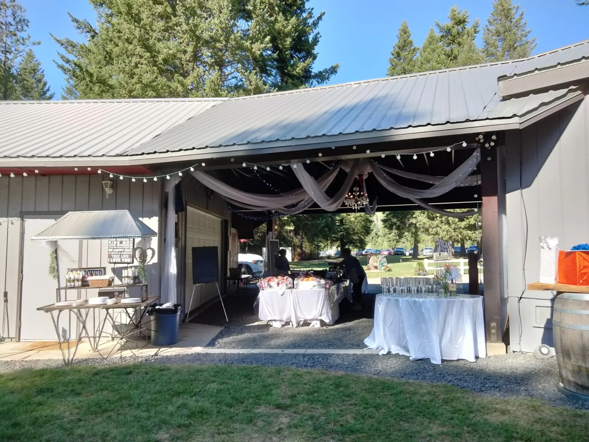 Outdoor event space with tables covered in white tablecloths, decorated with string lights, draped fabric, and a chandelier, set under a garage-like structure with trees and parked cars in the background.