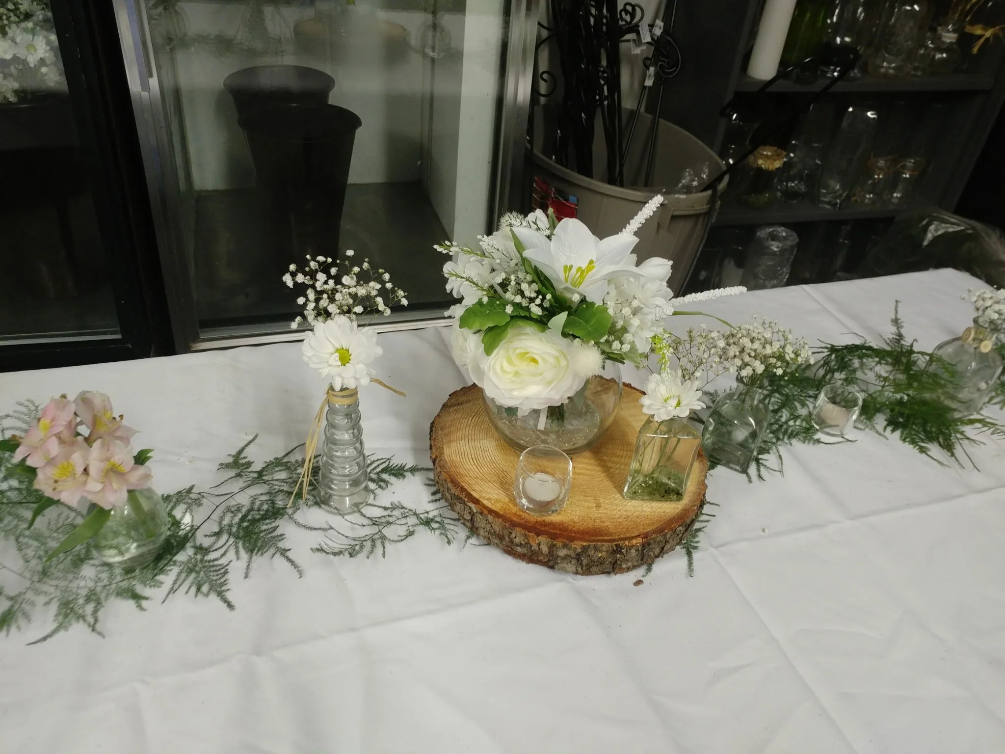 Floral table centerpiece with white flowers in glass vases, placed on a wooden slab, with greenery and small candles, set on a white tablecloth, in front of a window.