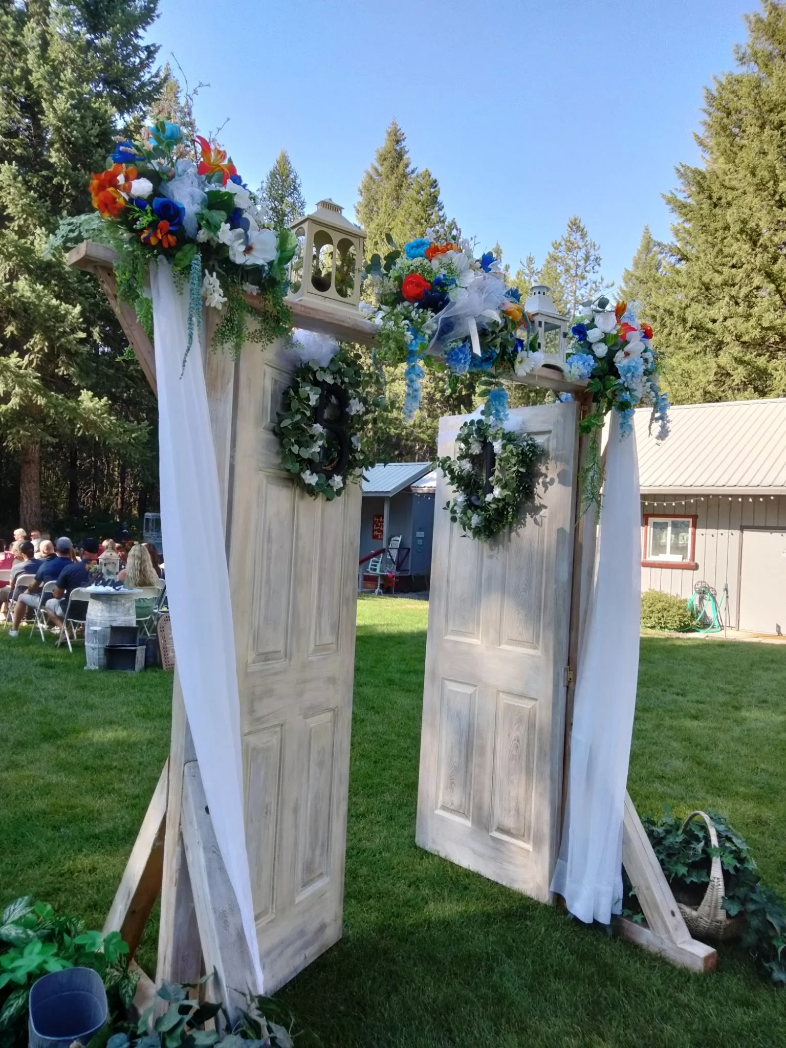 Wooden double doors decorated with floral arrangements, lanterns, and greenery, set up outdoors on grassy ground for a wedding or event.