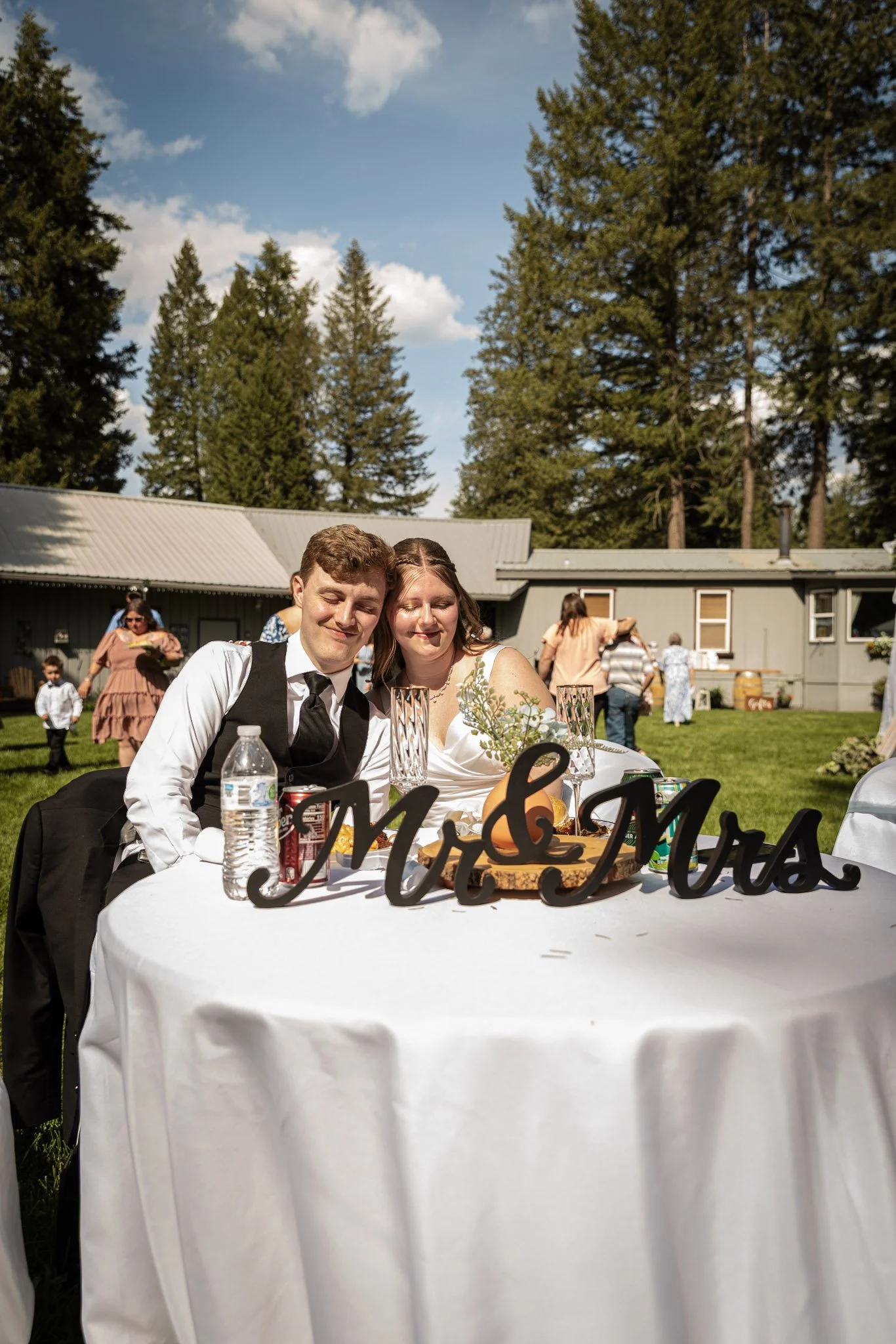 A bride and groom sit at a decorated table outside during a celebration, with a sign that partially reads 'Mr & Mrs'. The bride has long brown hair and is dressed in a white wedding gown, while the groom has short hair and is wearing a black vest, wh