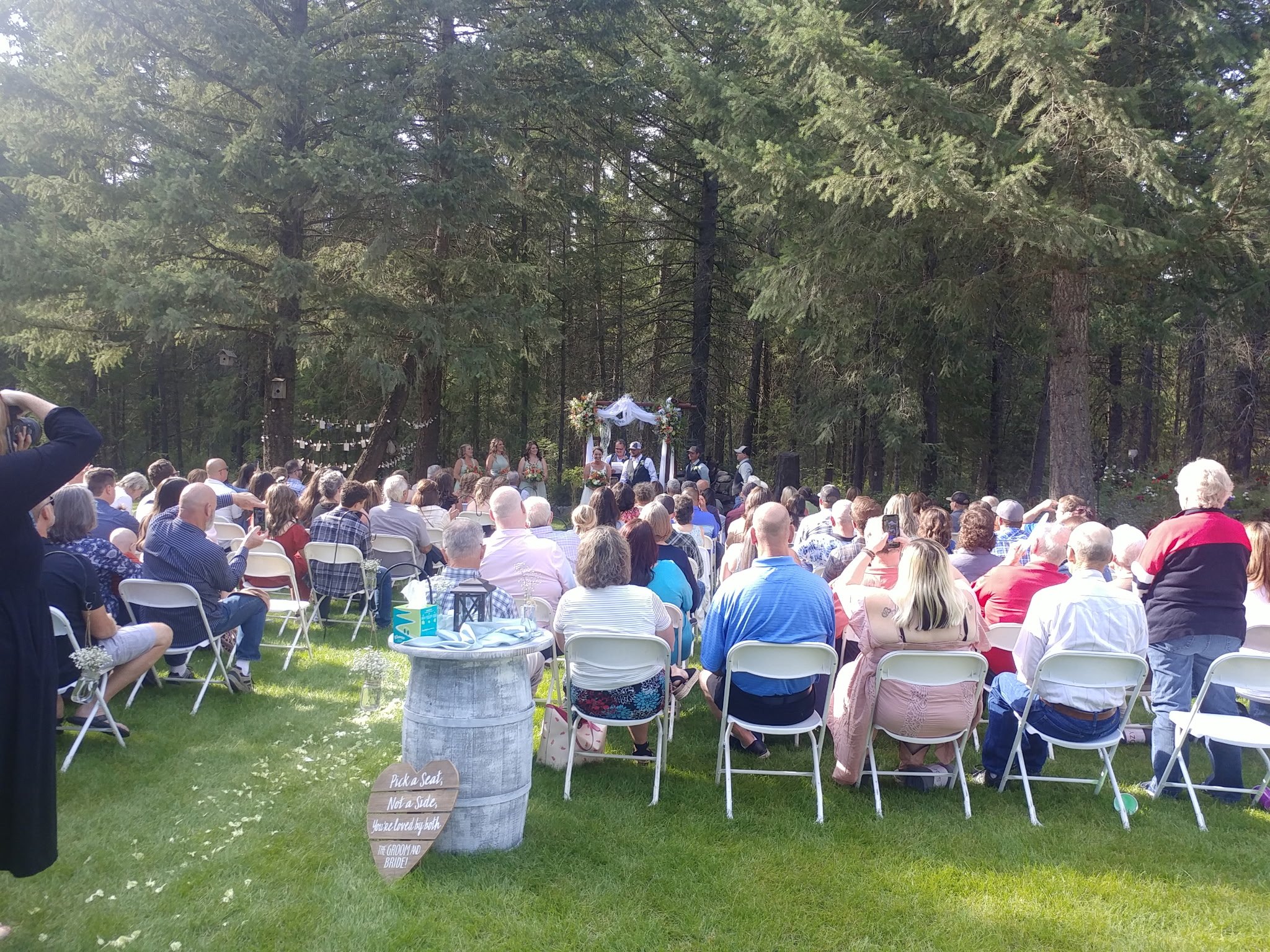 Outdoor wedding ceremony with guests seated on white chairs on grass, facing an arch decorated with flowers and white fabric, in a wooded area.