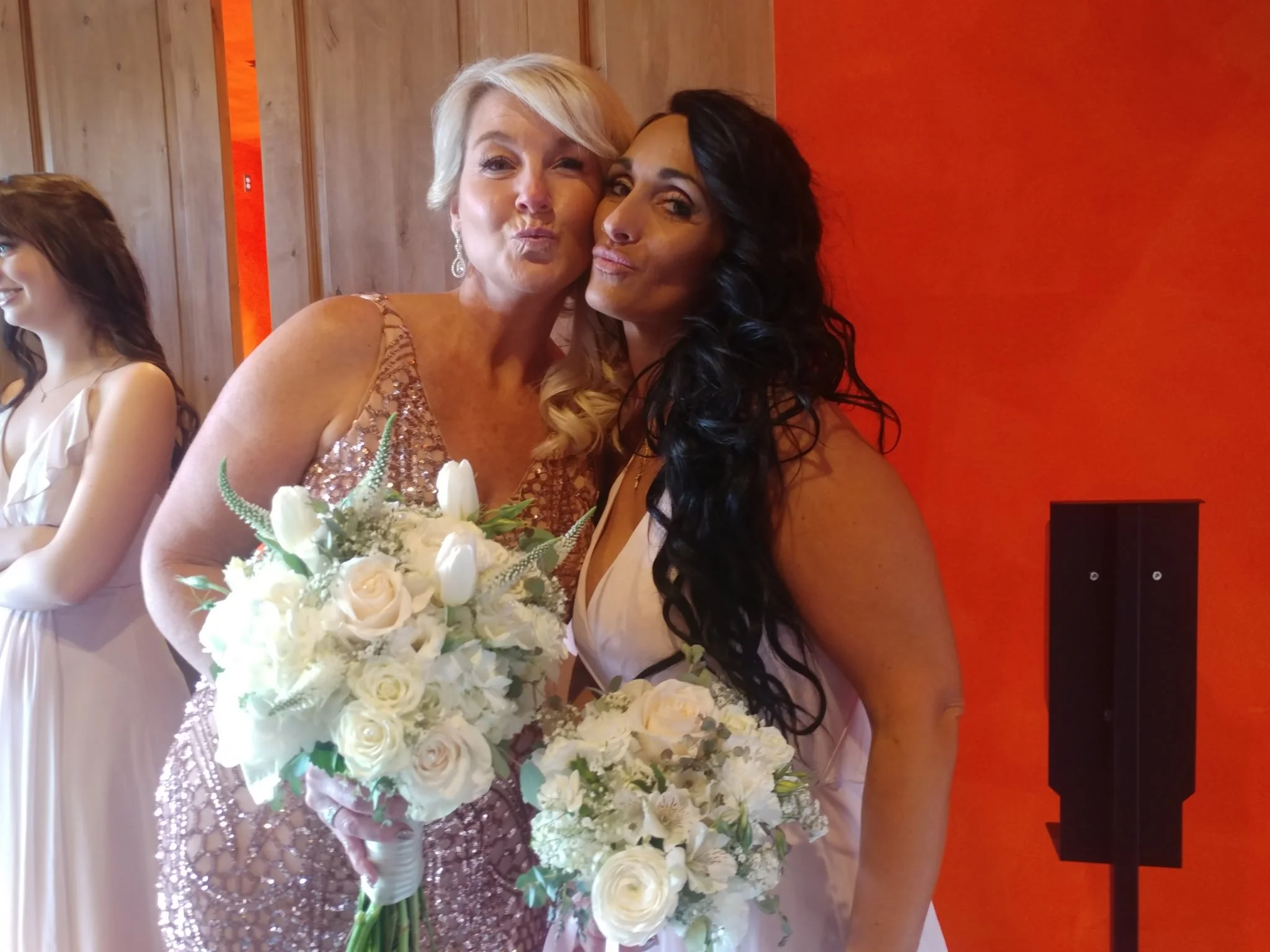 Two women at a wedding, one holding a bouquet of white flowers, posing together and making kiss faces. The woman on the left has short, platinum blonde hair and is wearing a sparkly, peach-colored dress. The woman on the right has long, dark curly ha