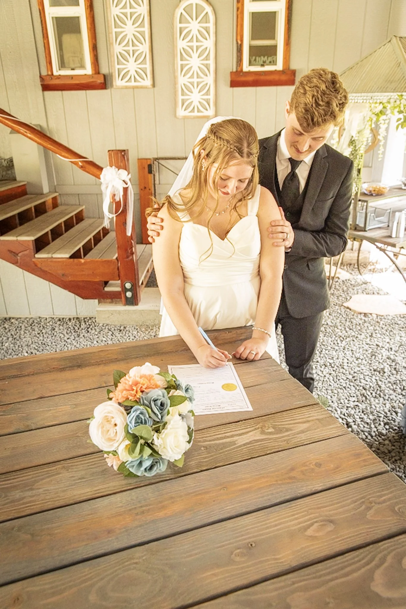 A bride and groom signing documents at a wedding ceremony, with a floral bouquet on a wooden table in the foreground.