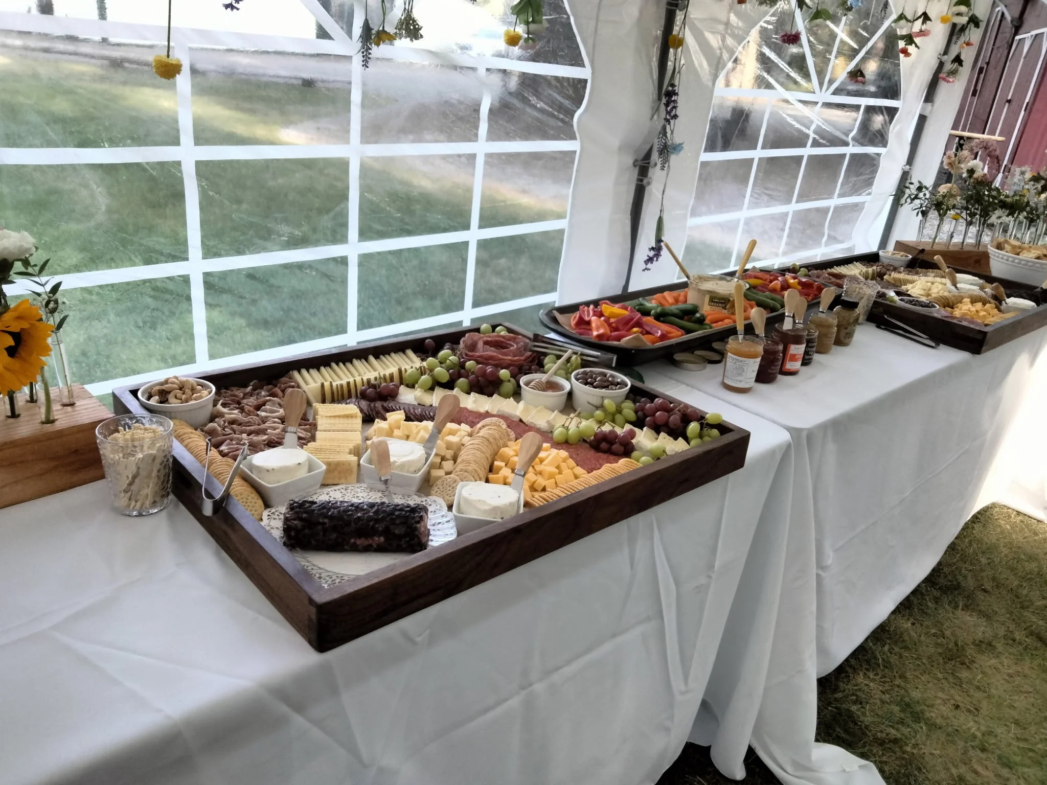 A cheese and charcuterie board spread on a table with fruit, crackers, nuts, and dips, set up inside a white tent with windows and greenery outside.