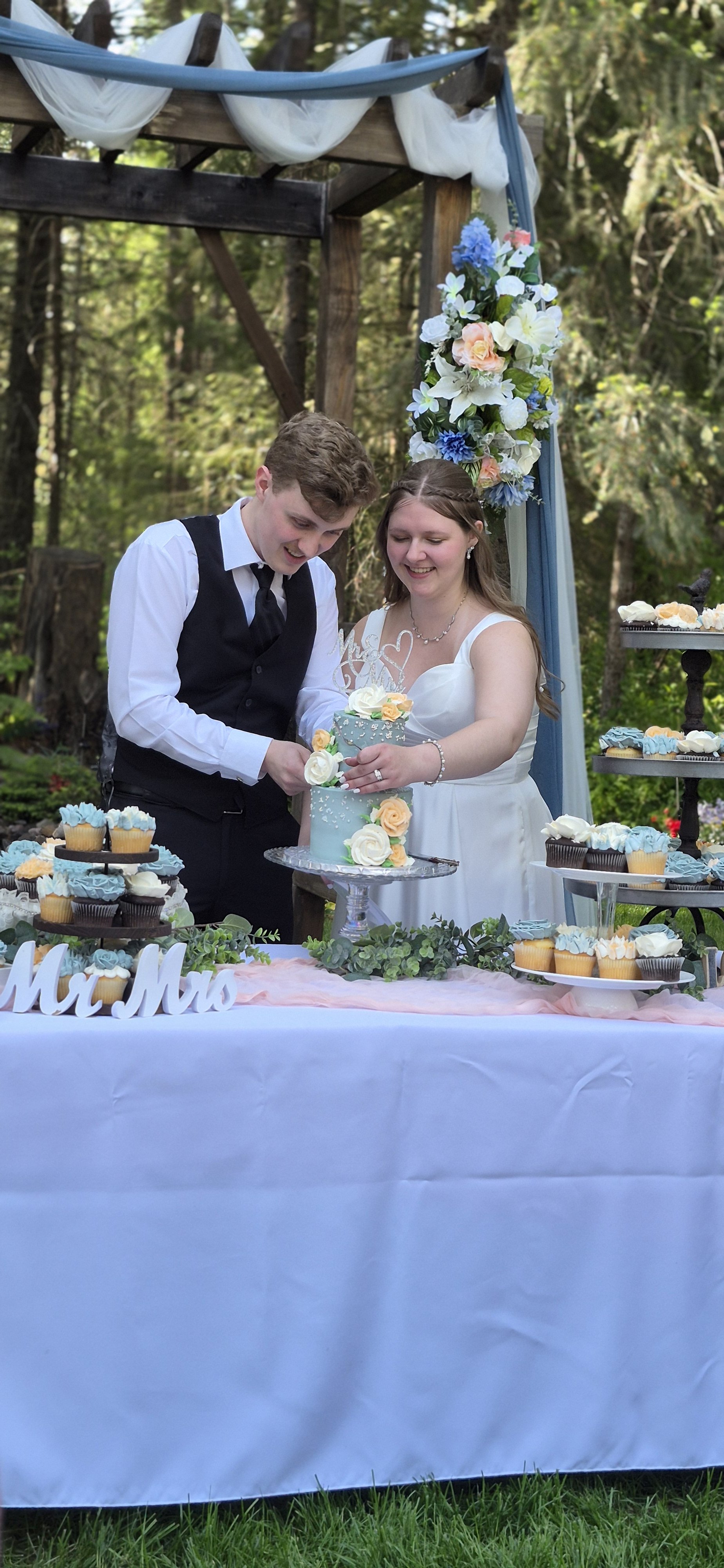 A couple in wedding attire cutting a wedding cake outdoors underneath a wooden arch decorated with white and blue fabric and flowers. The table has cupcakes and a "Mr & Mrs" sign, and is covered with a white tablecloth. They are smiling, surrounded b