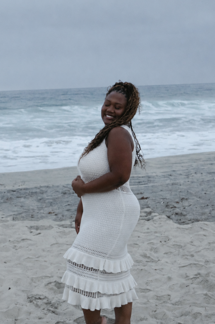 A woman in a white dress standing on the beach with the ocean and waves behind her, smiling with eyes closed.