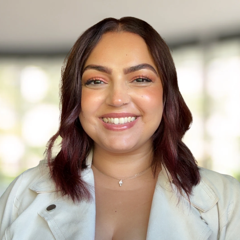 A young woman with shoulder-length brown hair and a big smile, wearing a light-colored blazer and a delicate necklace, standing indoors.