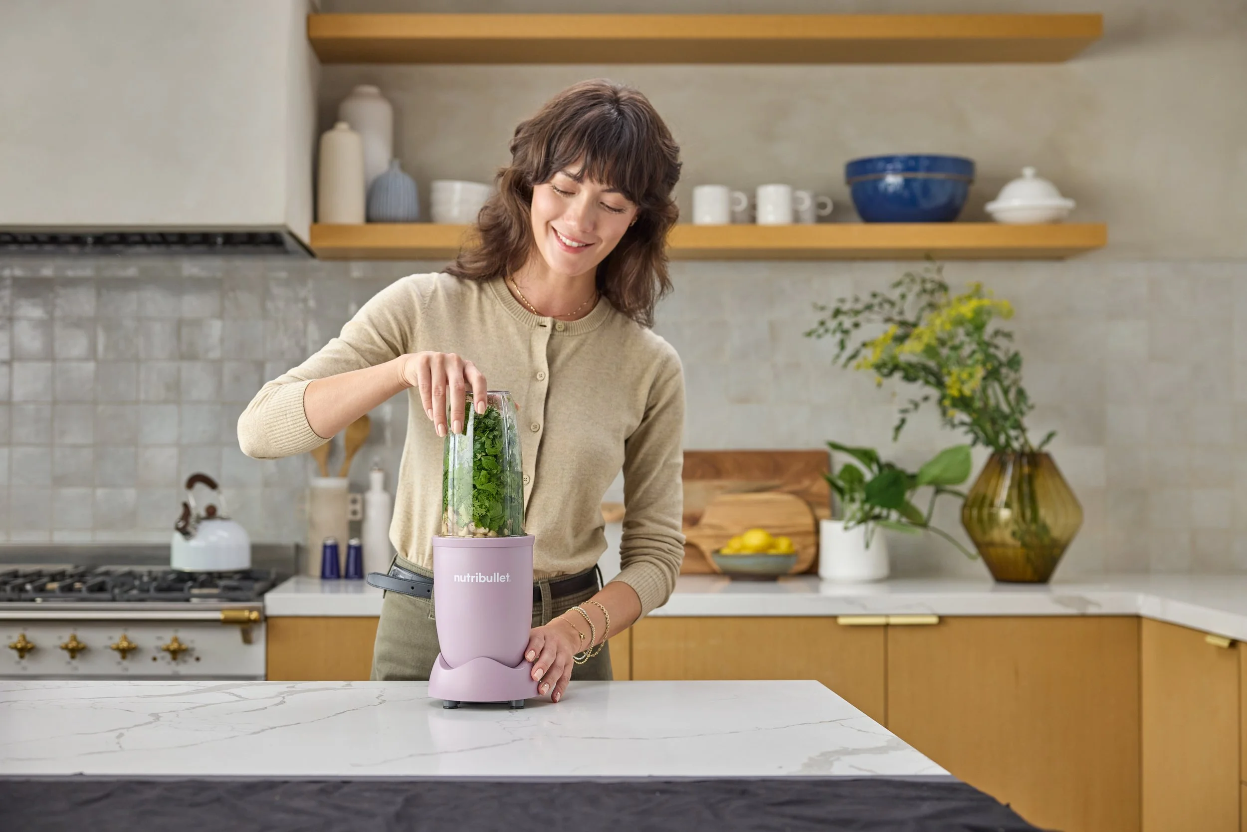 A woman smiling and preparing a smoothie in a pink NutriBullet blender in a modern kitchen with wooden cabinets, shelves with dishes, and a vase with flowers.