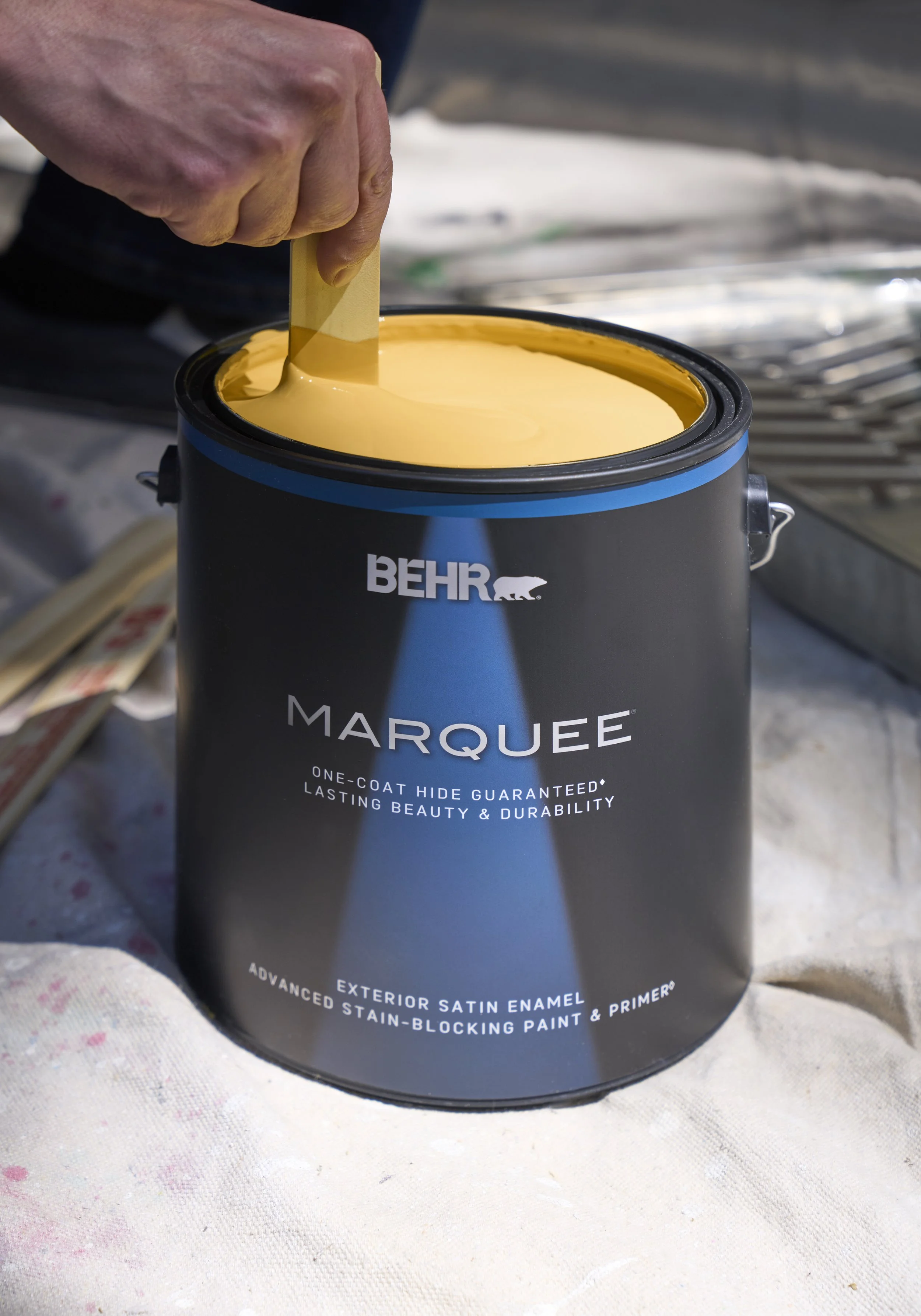 A hand dipping a paint stirrer into a can of beige exterior satin enamel paint labeled 'BEHR MARQUEE'.