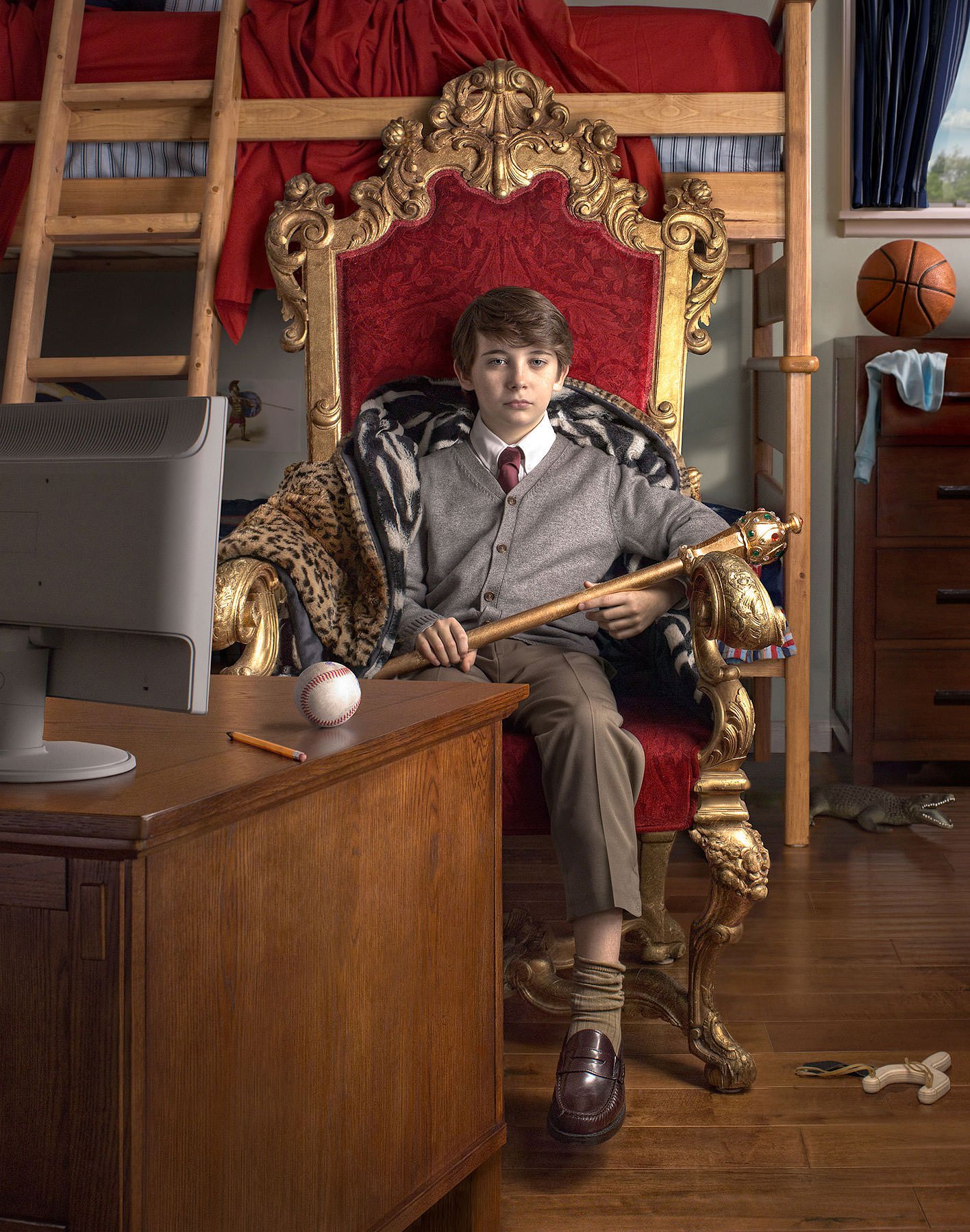 A young boy dressed in formal attire sitting on an ornate, gold-and-red throne-style chair in a room with a bunk bed. He is holding a golden scepter, and there is a baseball on a desk in front of him. The room has various items including a basketball