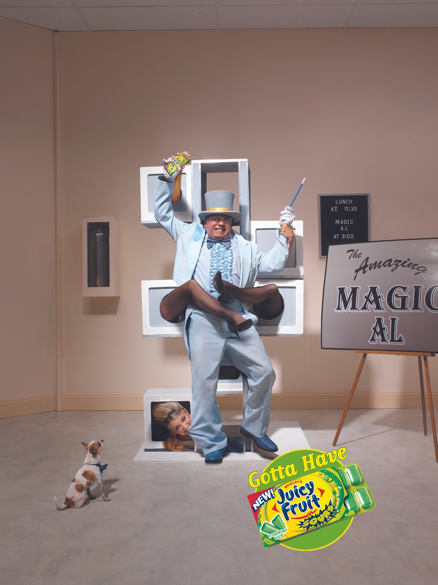 A man dressed as a magician in a light blue suit and top hat, holding a magic wand and a juice box, is standing inside a bookshelf with a woman’s head and legs sticking out from the bottom shelf. A small dog is sitting on the floor in front of him. T