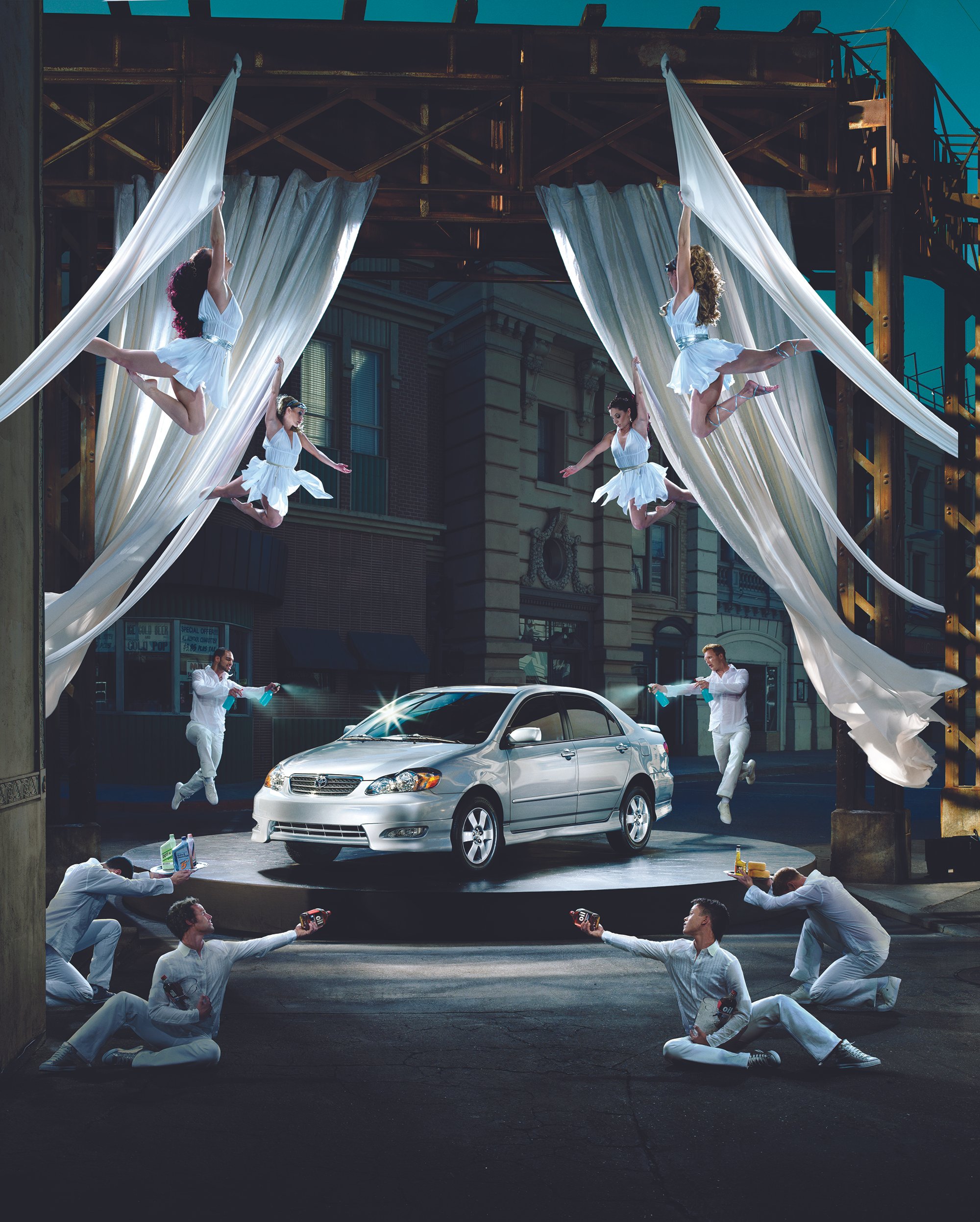 A city street with a theatrical car showcase featuring a silver sedan on a raised stage, surrounded by acrobatic performers and dancers dressed in white, with audience members in white sitting on the ground.
