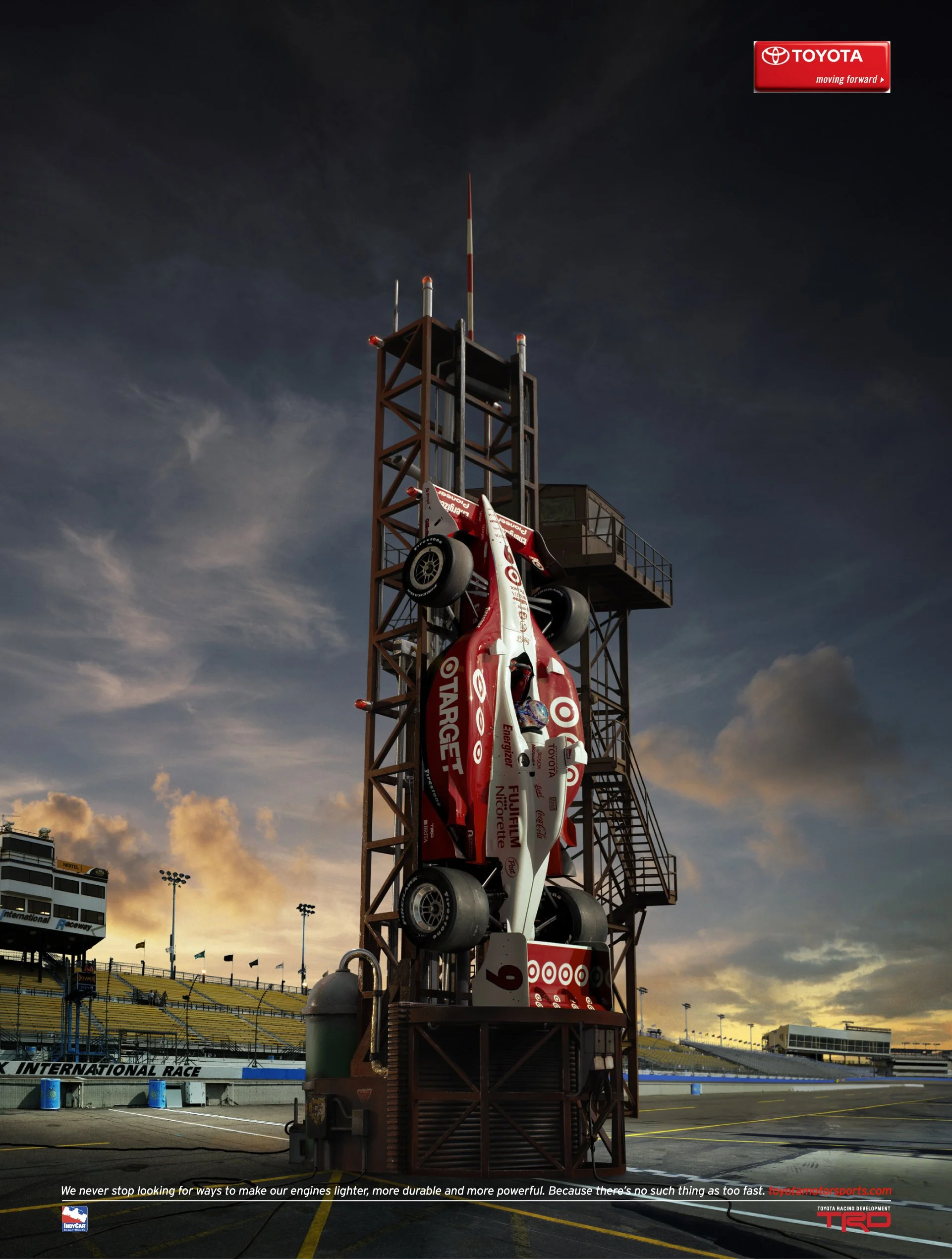 A racecar suspended vertically on a tall structure with stairs, outdoors at a race track during sunset, with the Toyota logo visible in the top right corner.