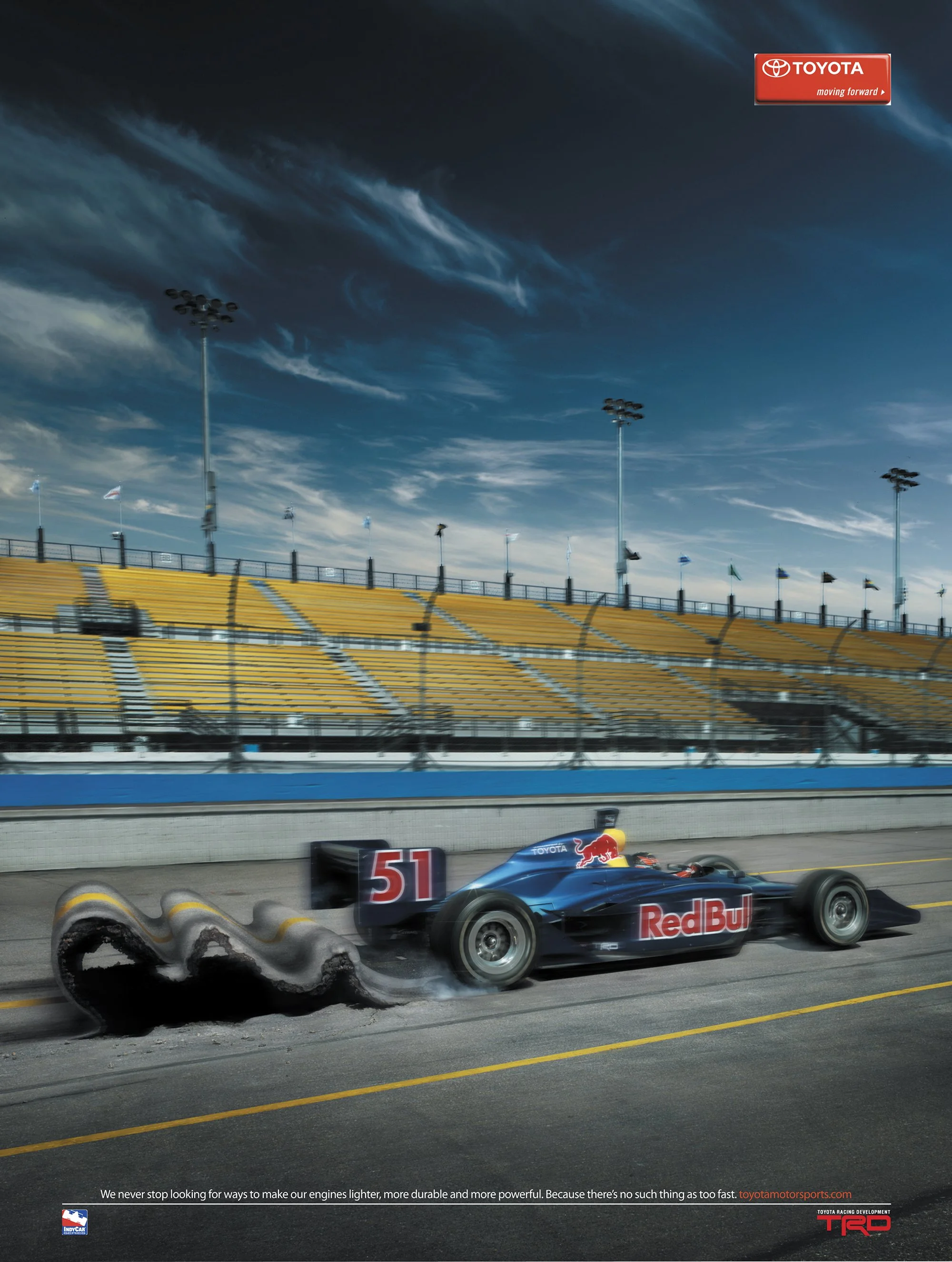 A blue race car with the number 51 on the side, emitting smoke from tire blowout, on a race track with yellow empty stands and a partly cloudy sky overhead. Toyota and Red Bull sponsorship logos are visible.