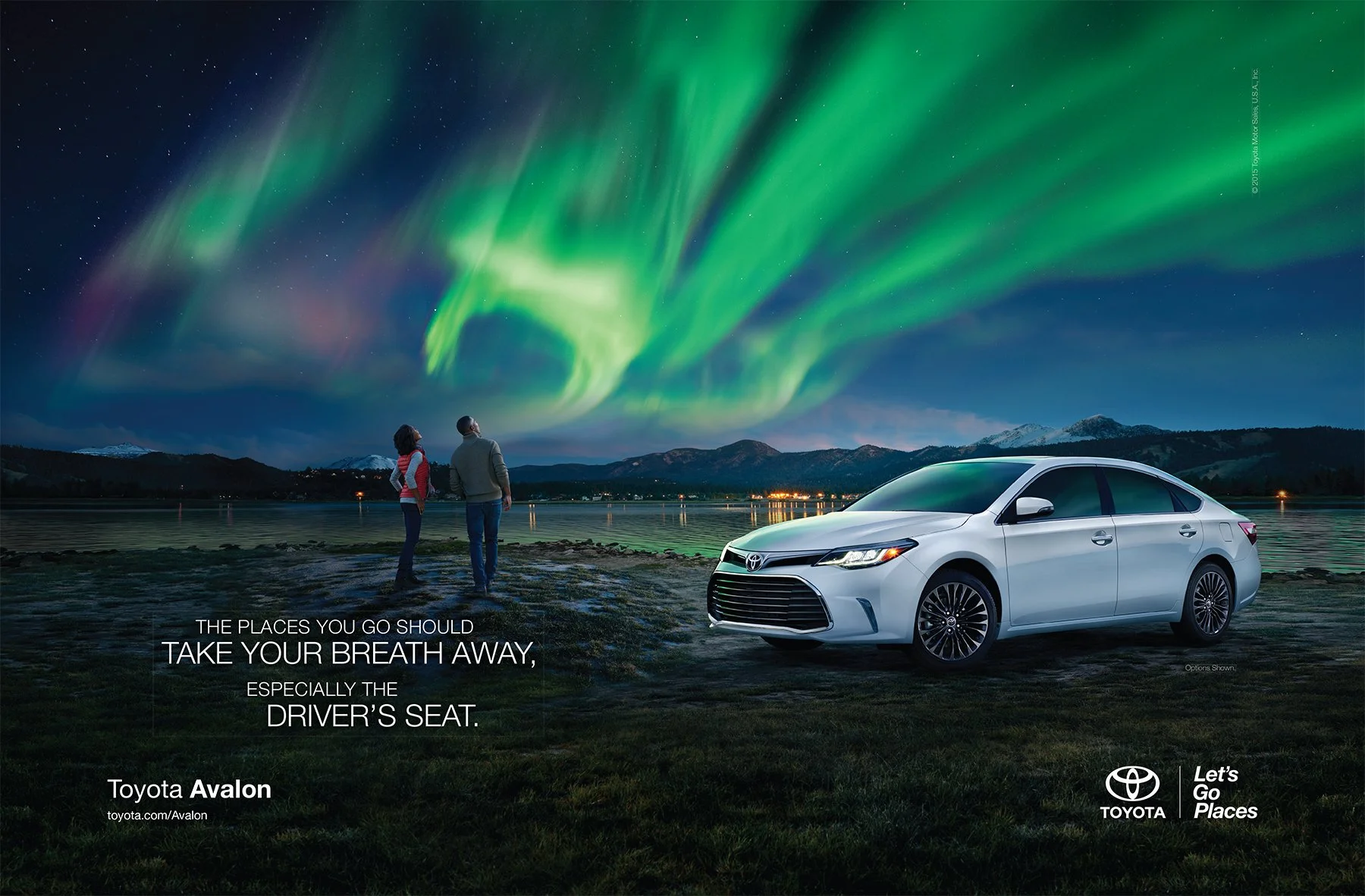 A Toyota car parked near a lakeshore with two people looking at the northern lights in the sky over mountains.
