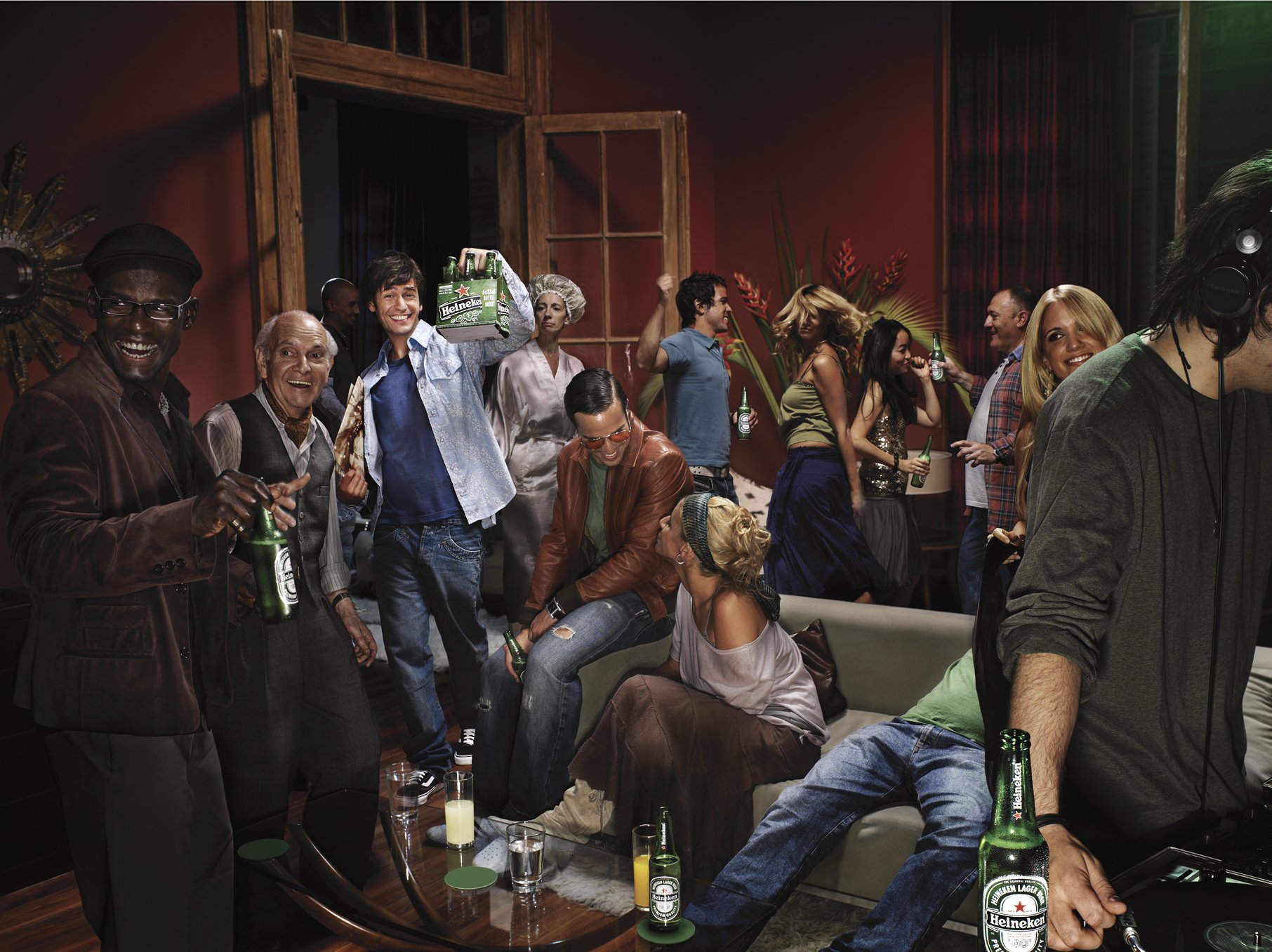 People dancing and socializing at a party, holding drinks, with a DJ playing music on the right side.