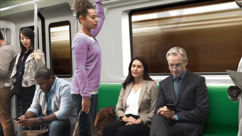 A woman standing in a purple sweater is holding onto the overhead handrail while a man and a woman seated on a subway train are looking at their phones, and two women are standing in the background.