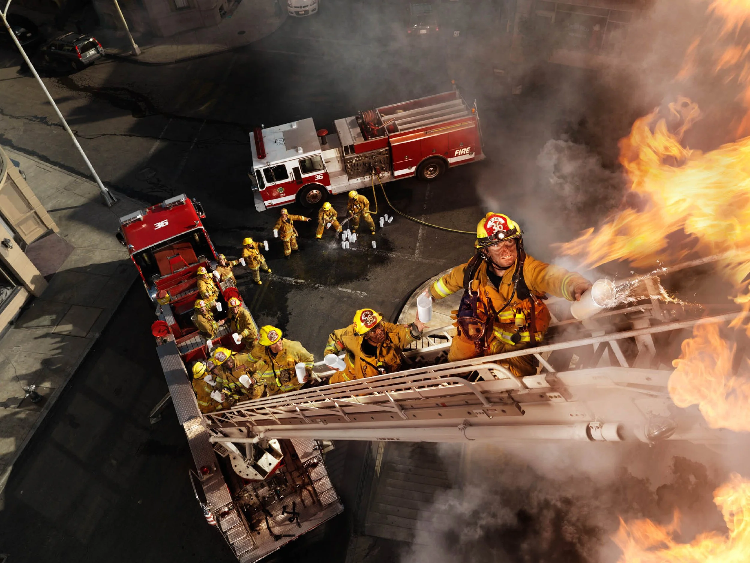 Firefighters battling a blaze on a building fire ladder at night, with fire trucks and firefighters in yellow gear on the street.