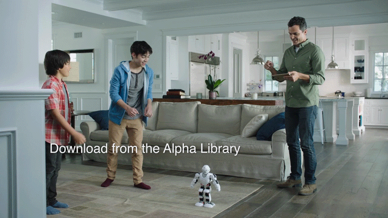 A man and two boys are in a living room with a robot on the floor. The man is holding a tablet and smiling, while the boys look at the robot. The room has a sofa, a table with books, and a kitchen in the background.