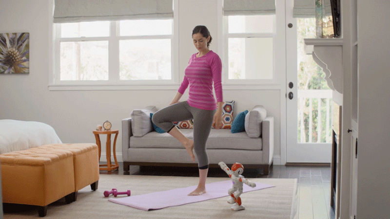 A woman in a pink and purple striped sweater and gray leggings practices yoga on a pink mat in a bright living room, with a small robotic dog toy nearby.