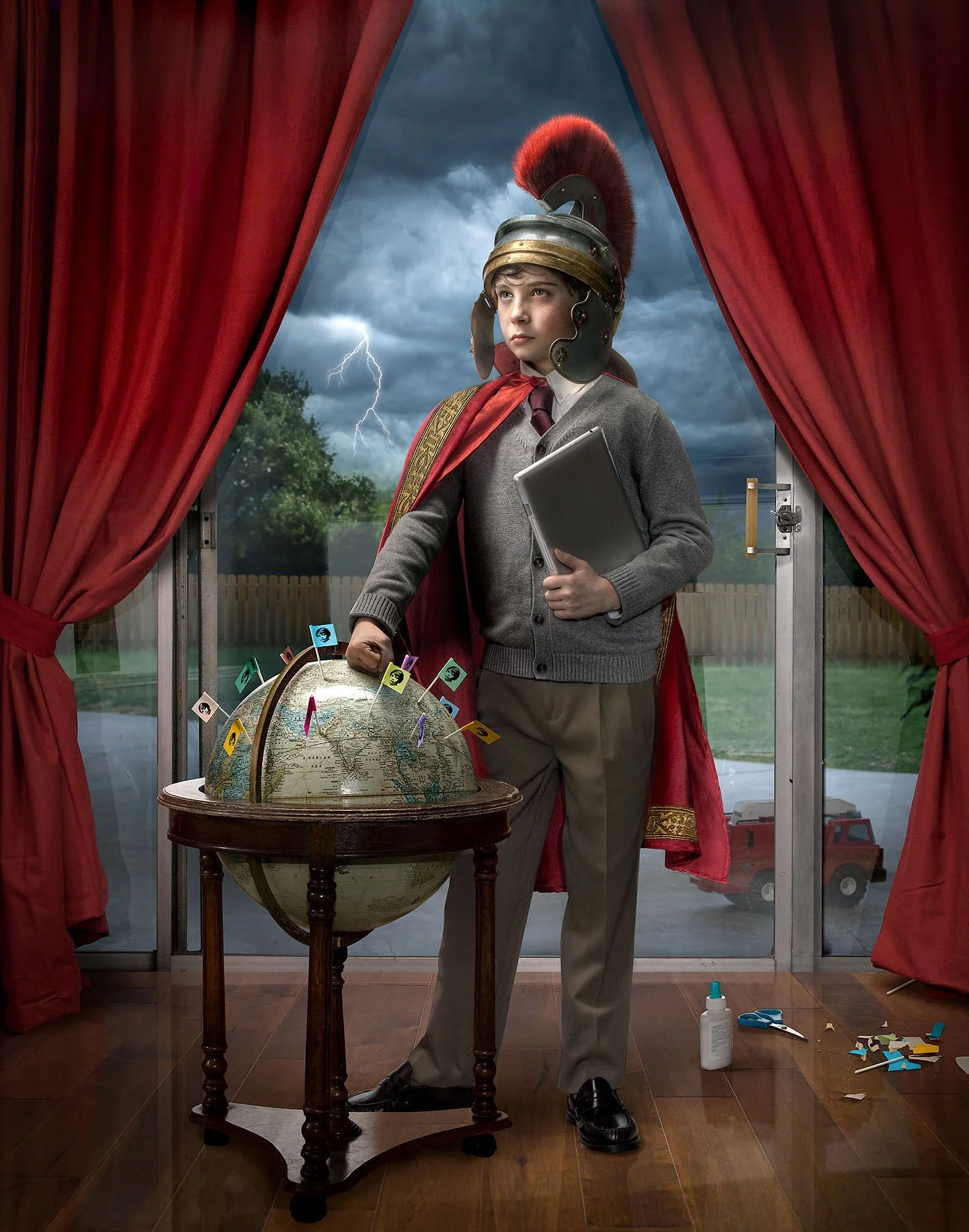 A young boy dressed as a Roman senator or general, wearing a helmet with a red plume, standing in front of a window with red curtains. He holds a laptop in one hand and is pointing to a large globe on a table with small flags on it. Outside the windo
