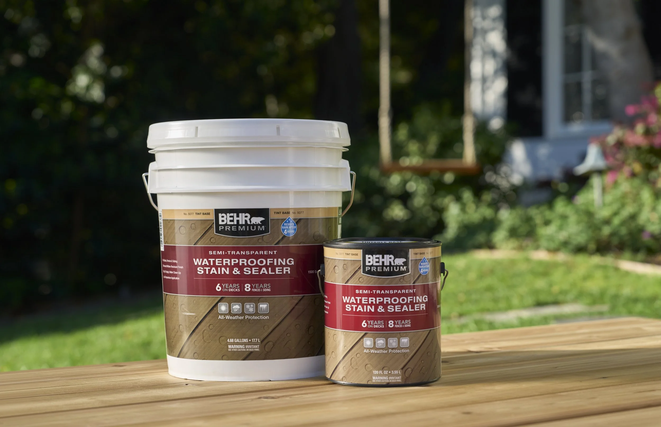 Two containers of BEHR Premium Waterproofing Stain & Sealer, with a larger white bucket and a smaller metal can, placed on a wooden surface outdoors with a blurred background of grass, trees, and a house with a porch