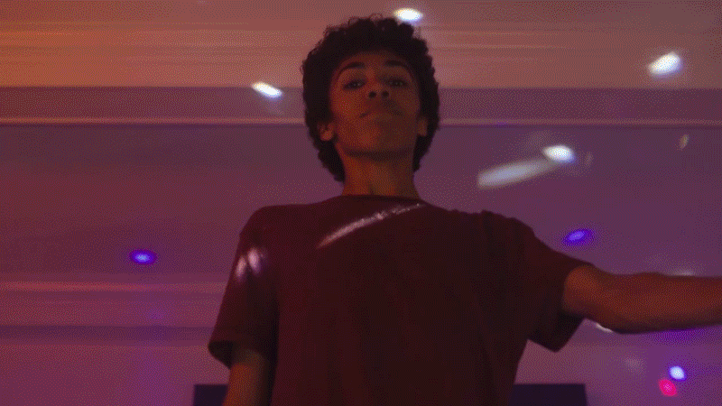 Person with short curly hair standing in a room with purple and blue lights, facing camera with a neutral expression.
