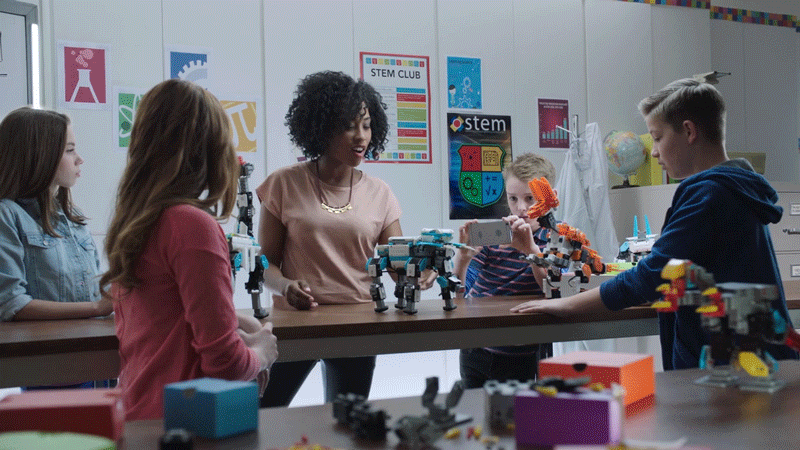 A teacher and four students in a classroom showcasing LEGO robots and models on the table.