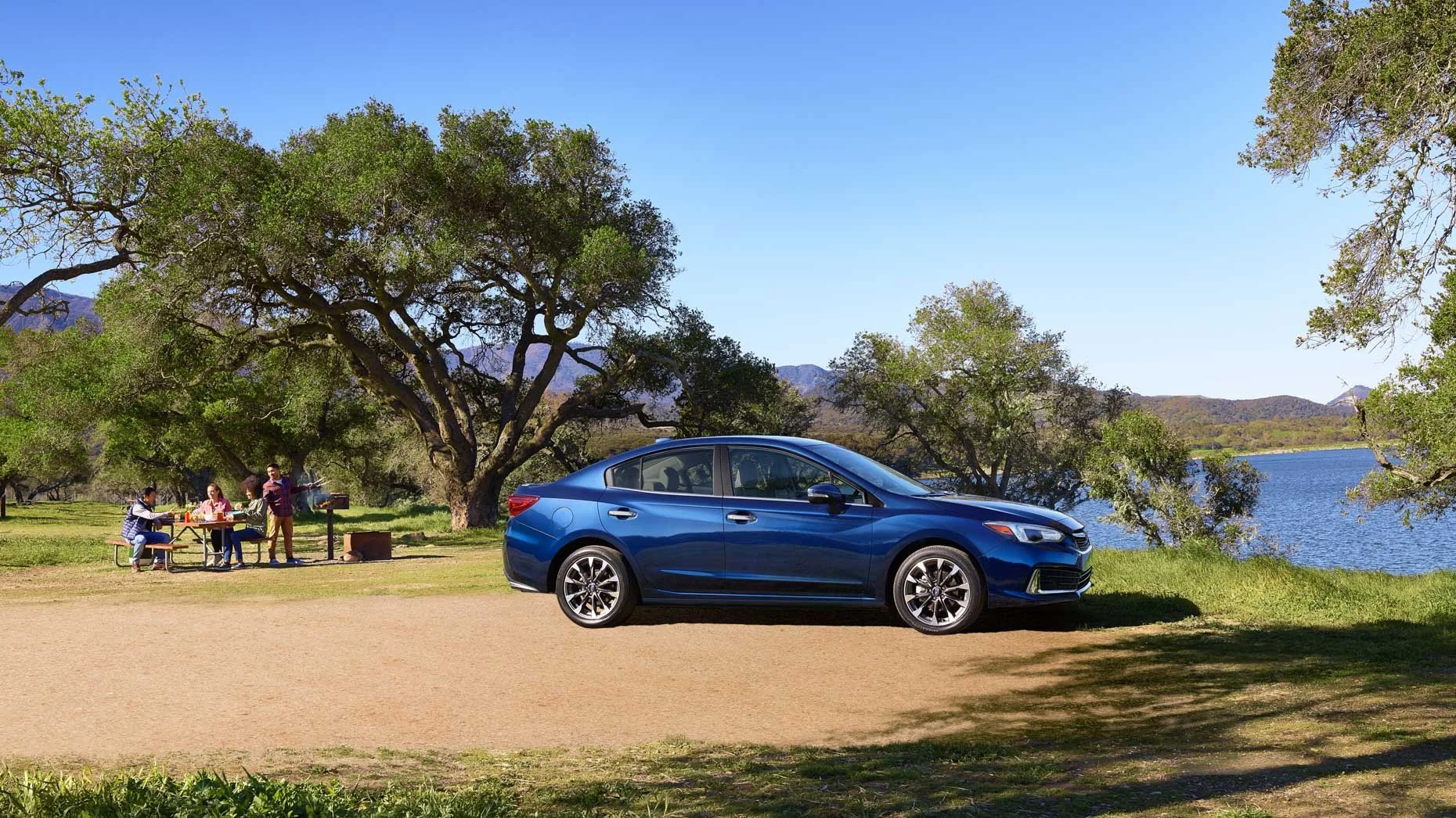 A blue sedan parked near a group of people having a barbecue picnic near a lake with trees and mountains in the background.
