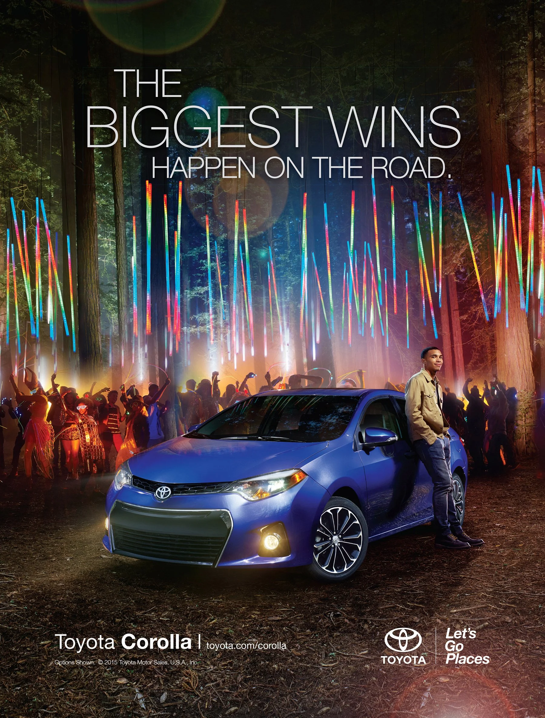 A blue Toyota Corolla parked on a dirt path in a forest at night with colorful light displays and a crowd of people dancing in the background. A man stands beside the car, smiling.