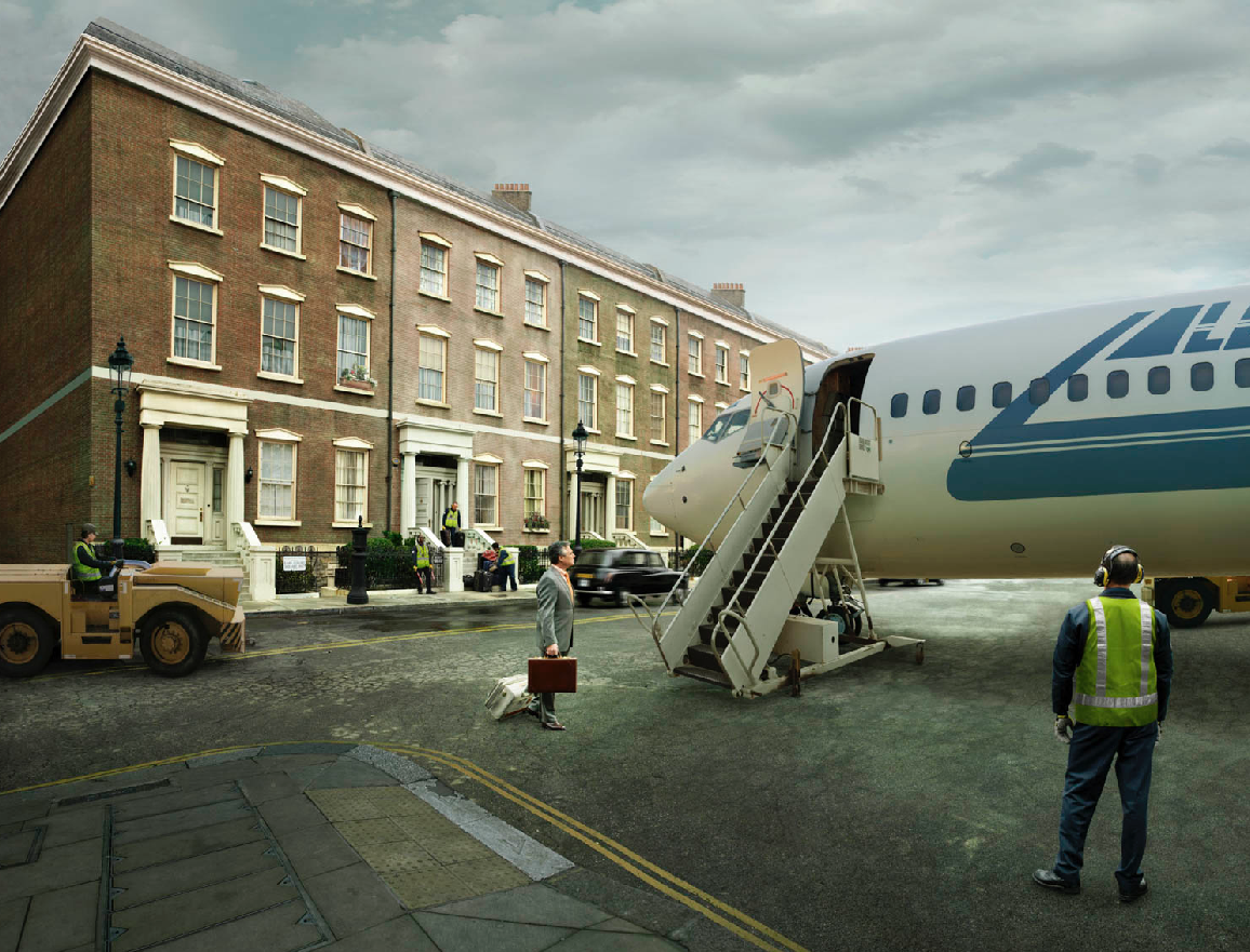 Business traveler in a suit with a briefcase arriving at a small regional airport, walking towards a small private jet, with airport staff and ground crew assisting. Residential buildings line the street in the background, under a cloudy sky.