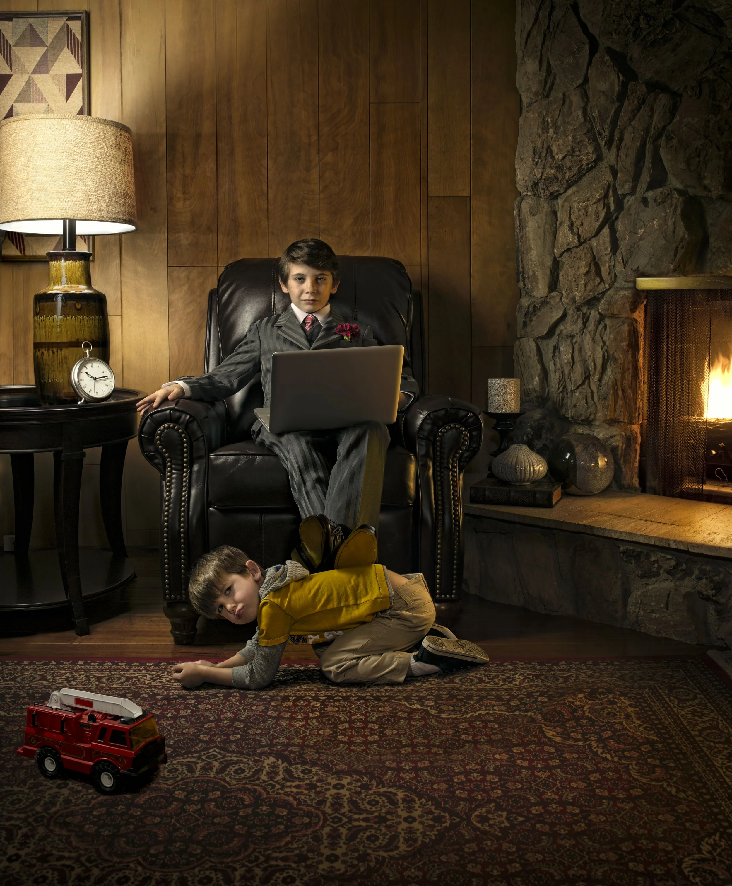 A young boy in a suit sitting with a laptop on an armchair beside a fireplace, and another child playing on the carpet with a toy fire truck.