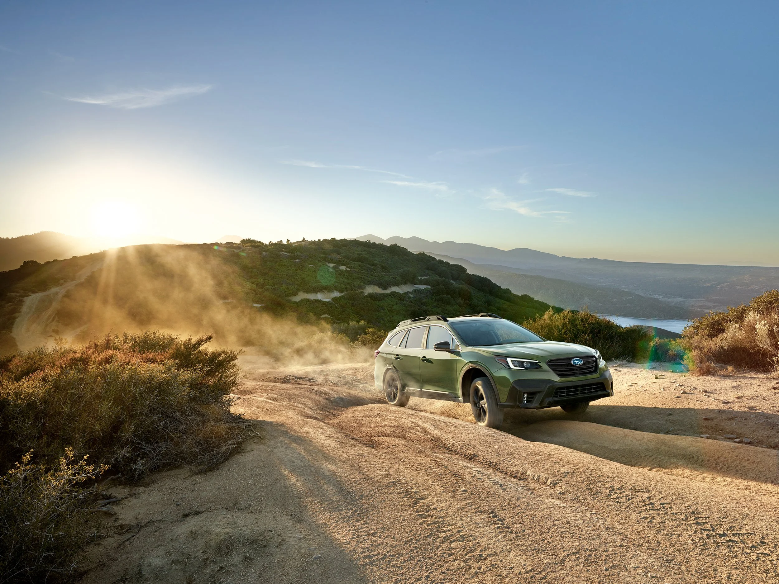 A green SUV driving on a dirt trail through a desert landscape with mountains in the distance and the sun setting on the horizon.