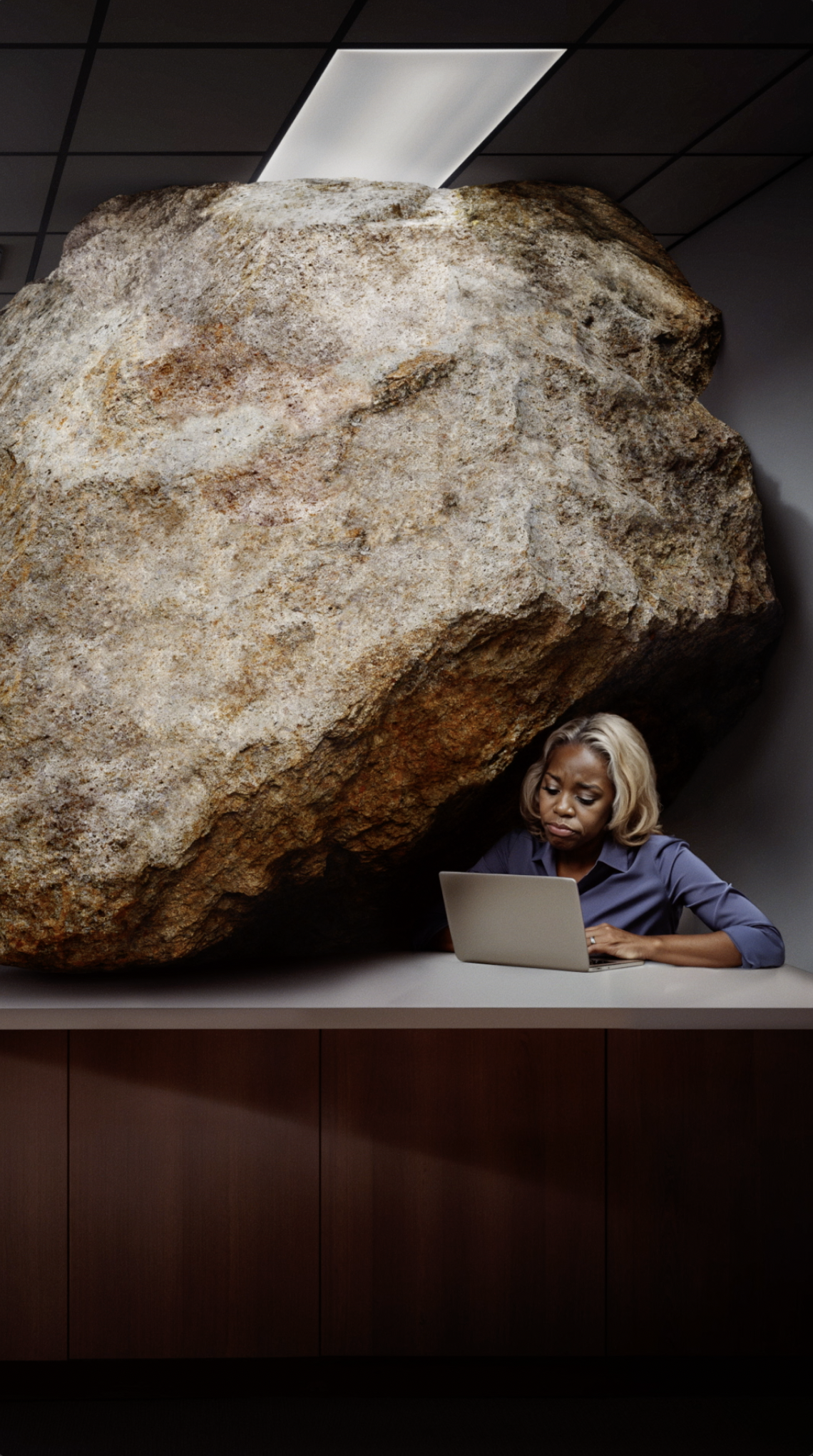 A woman working on a laptop at a desk with a large boulder behind her in an office setting.
