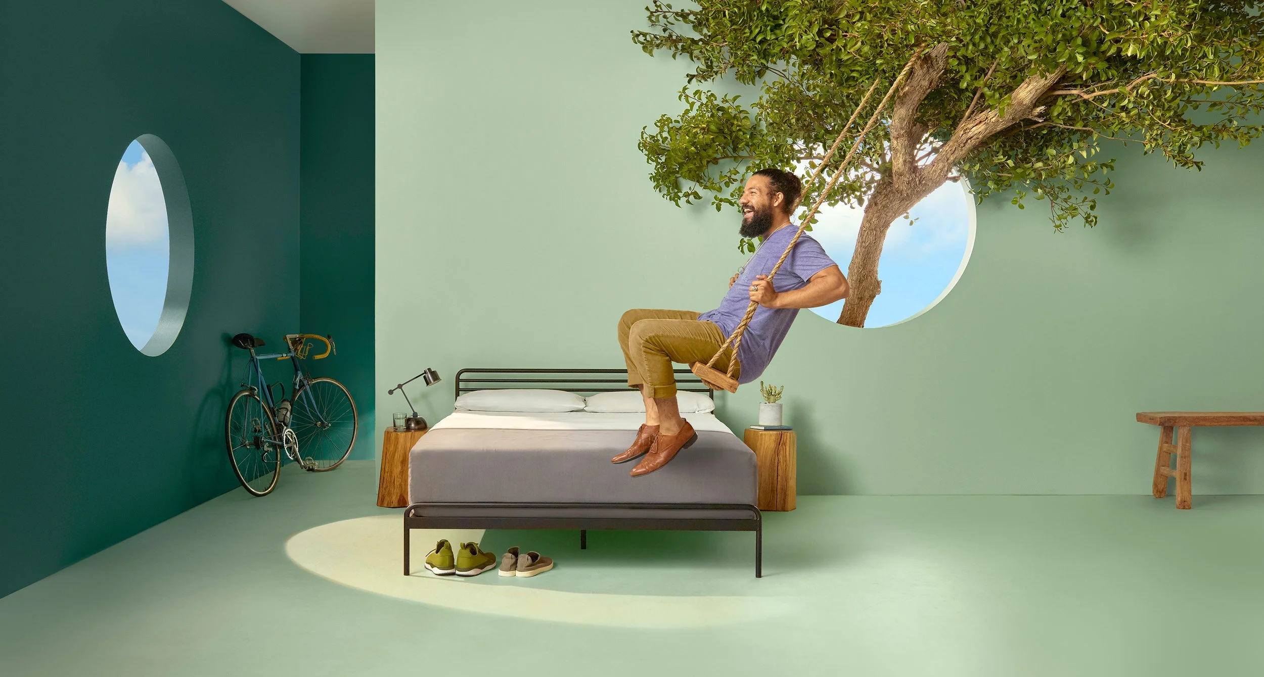 A man sitting on a swing attached to a tree that extends through a round window in a bedroom wall, with a bicycle and a small table with a lamp nearby, and shoes on the floor.