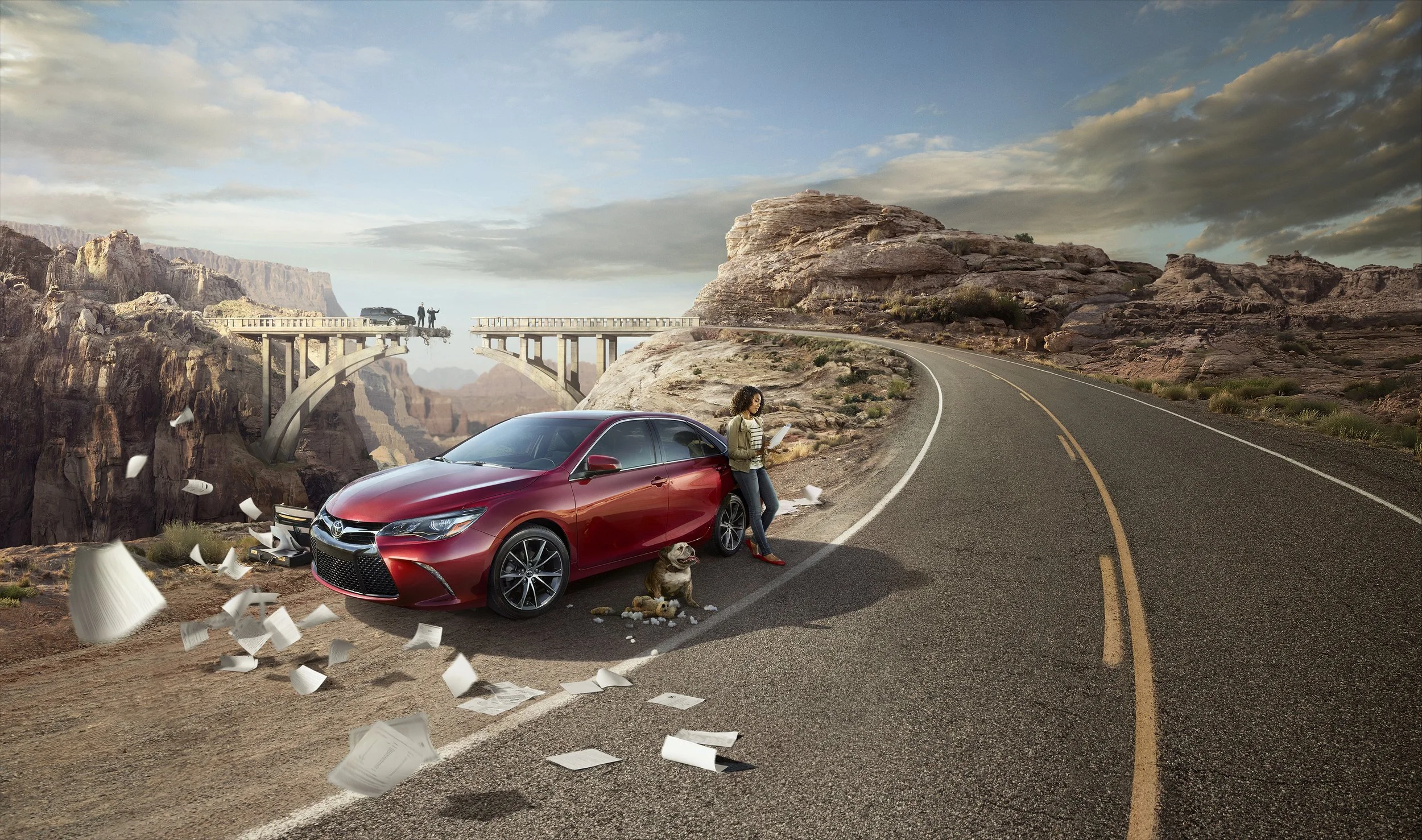 A woman with a dog standing beside a red car on a winding road in a canyon landscape with a bridge in the background.