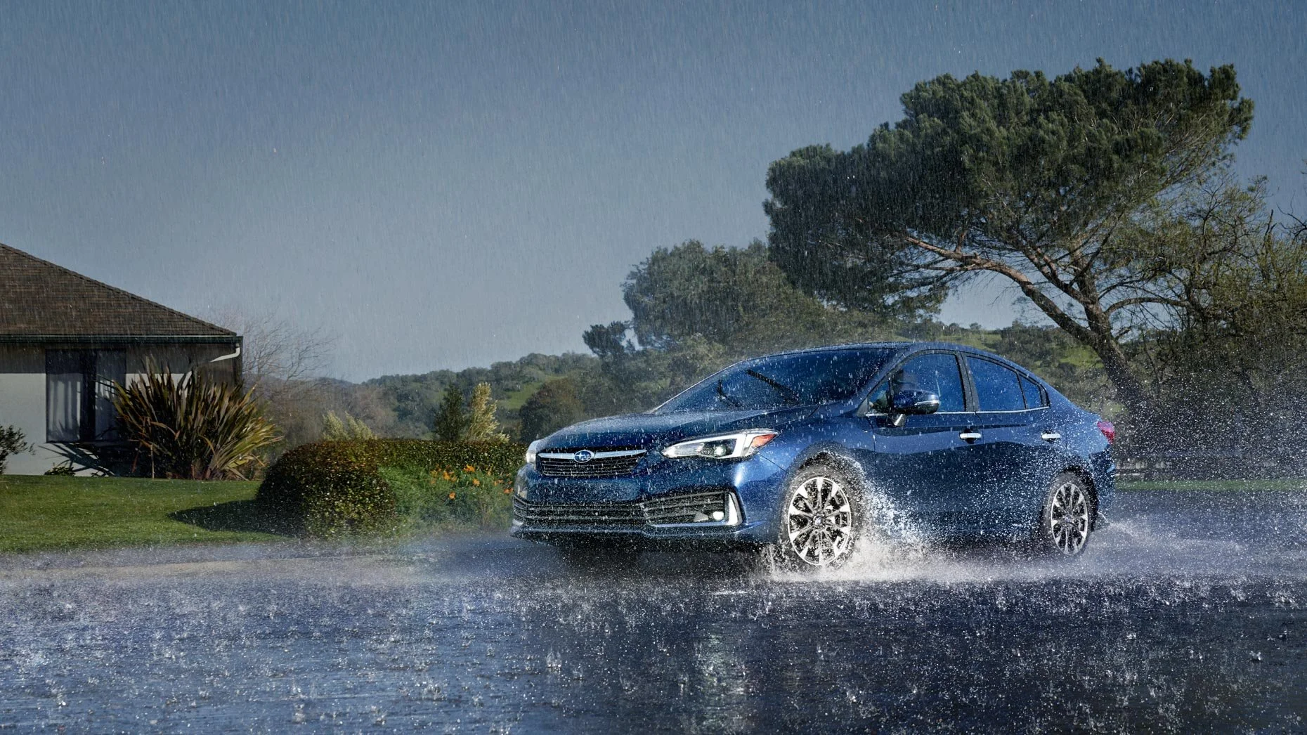 A blue Subaru sedan driving through a rainy street with water splashing around.