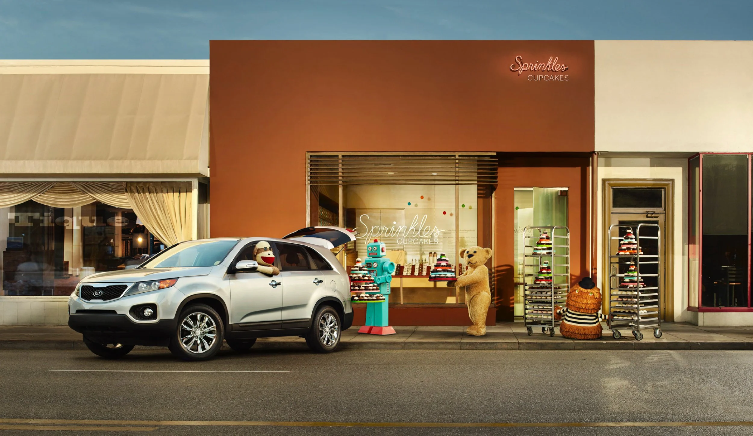 A storefront called Sprinkles Cupcakes with a silver SUV parked in front. Two stuffed animals, a monkey and a bear, stand outside the shop near displays of cupcakes. A blue robot figure holds cupcakes on a stand.