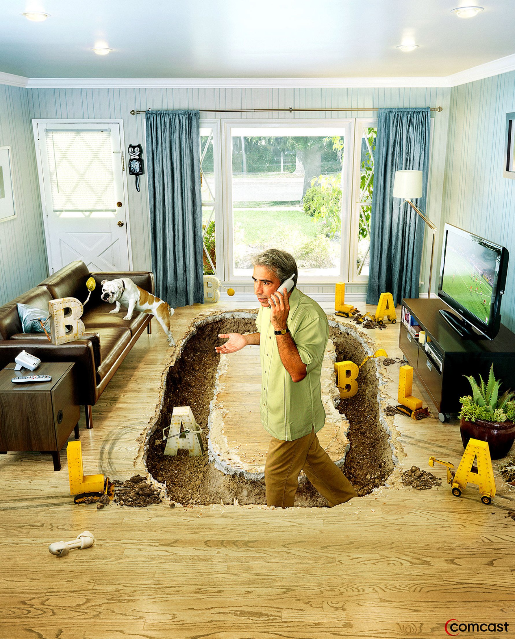 Man talking on the phone with a large hole in the center of a living room floor, surrounded by toys and block letters, with a dog on the couch and a television showing a soccer game.