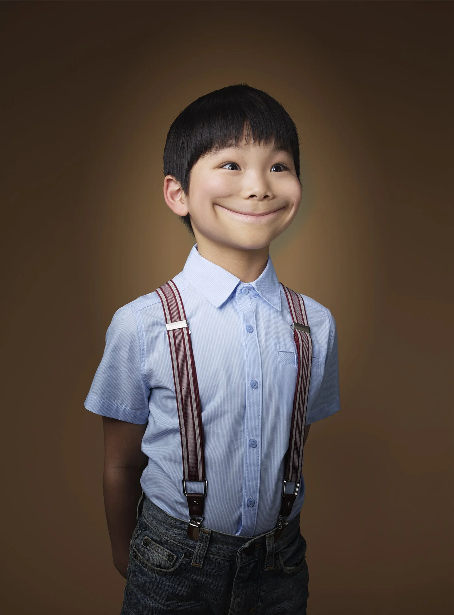 A young boy with black hair, wearing a light blue button-up shirt with suspenders, smiling with dimples, standing against a brown background.