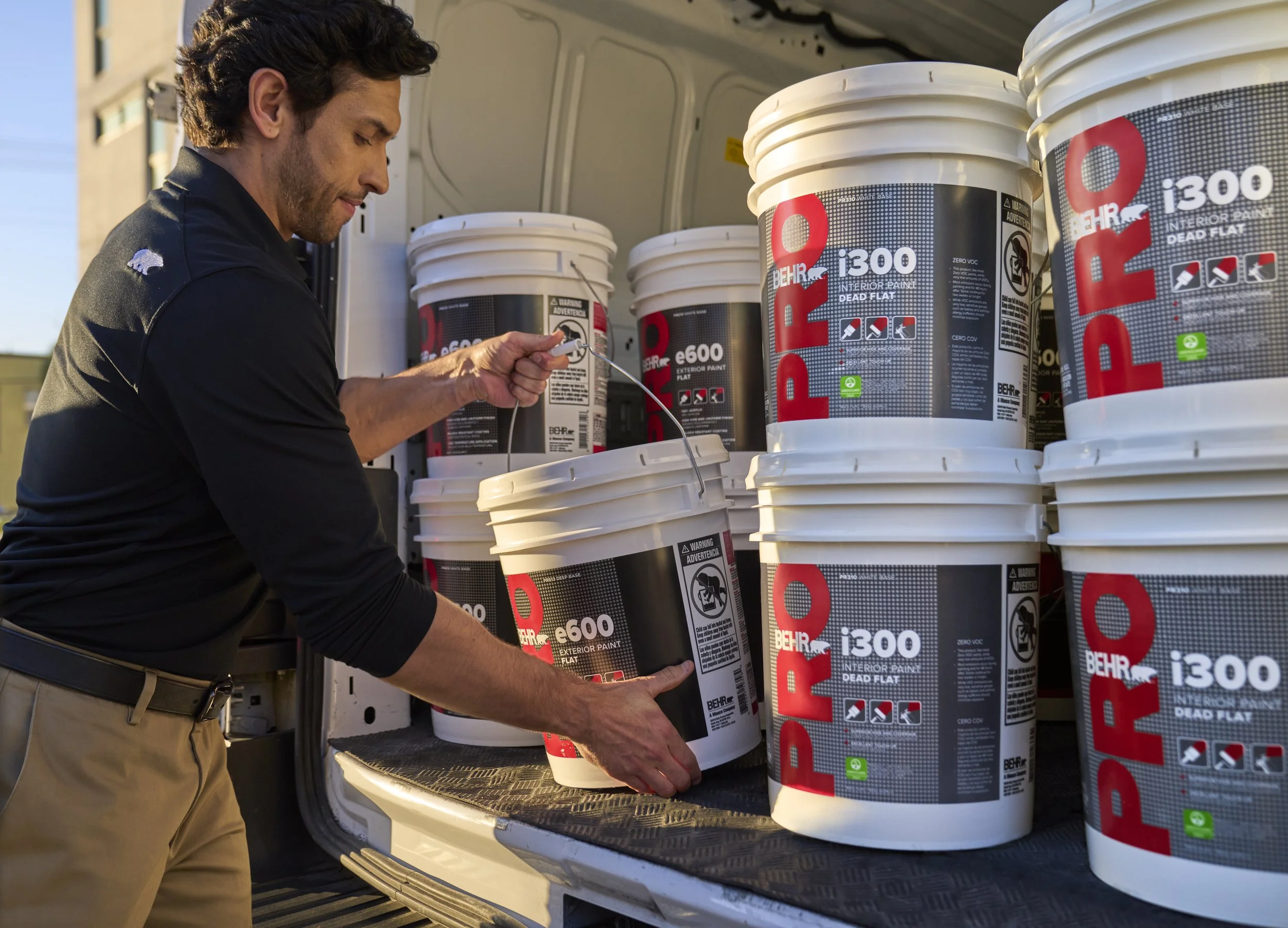 A man in black and khaki clothing is loading white buckets of BEHR interior and exterior paint into the back of a truck, with multiple paint buckets stacked around him.