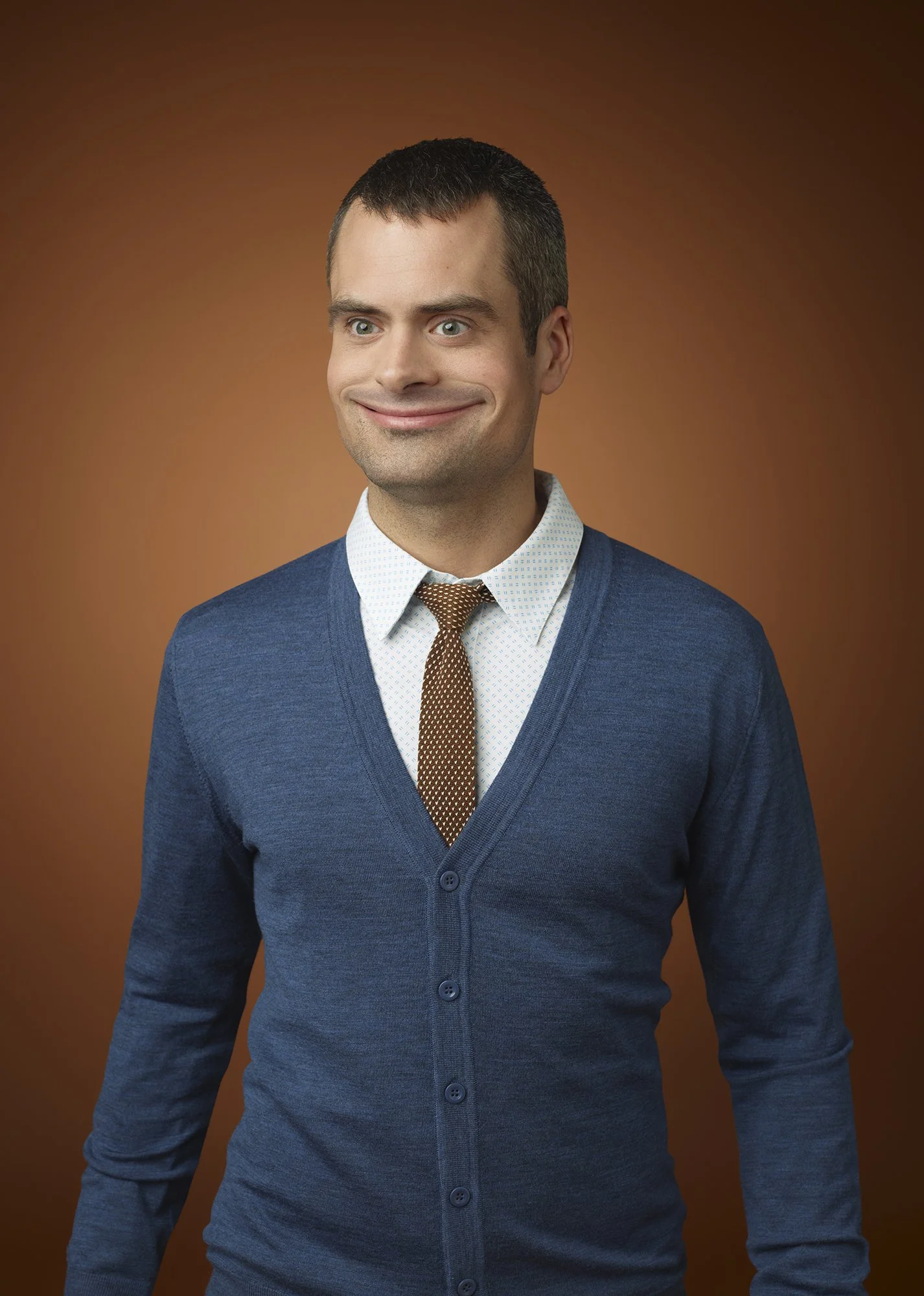 A man with short dark hair, wearing a blue cardigan, white dress shirt, and brown patterned tie, smiling subtly against a brown background.