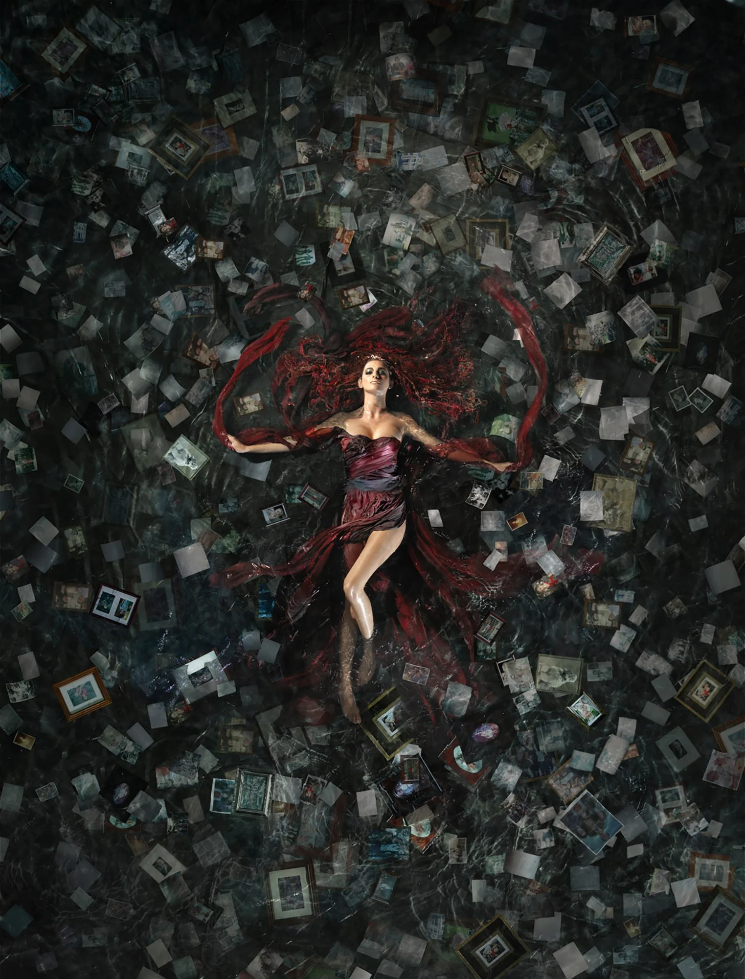 A woman in a dark red dress is lying on a black marble floor surrounded by scattered photo frames and photographs, her red hair spread out around her.