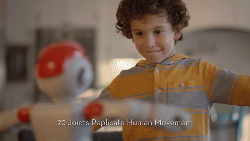 A young boy smiling as he interacts with a robot that has a spherical head and animated arms, in an indoor setting.