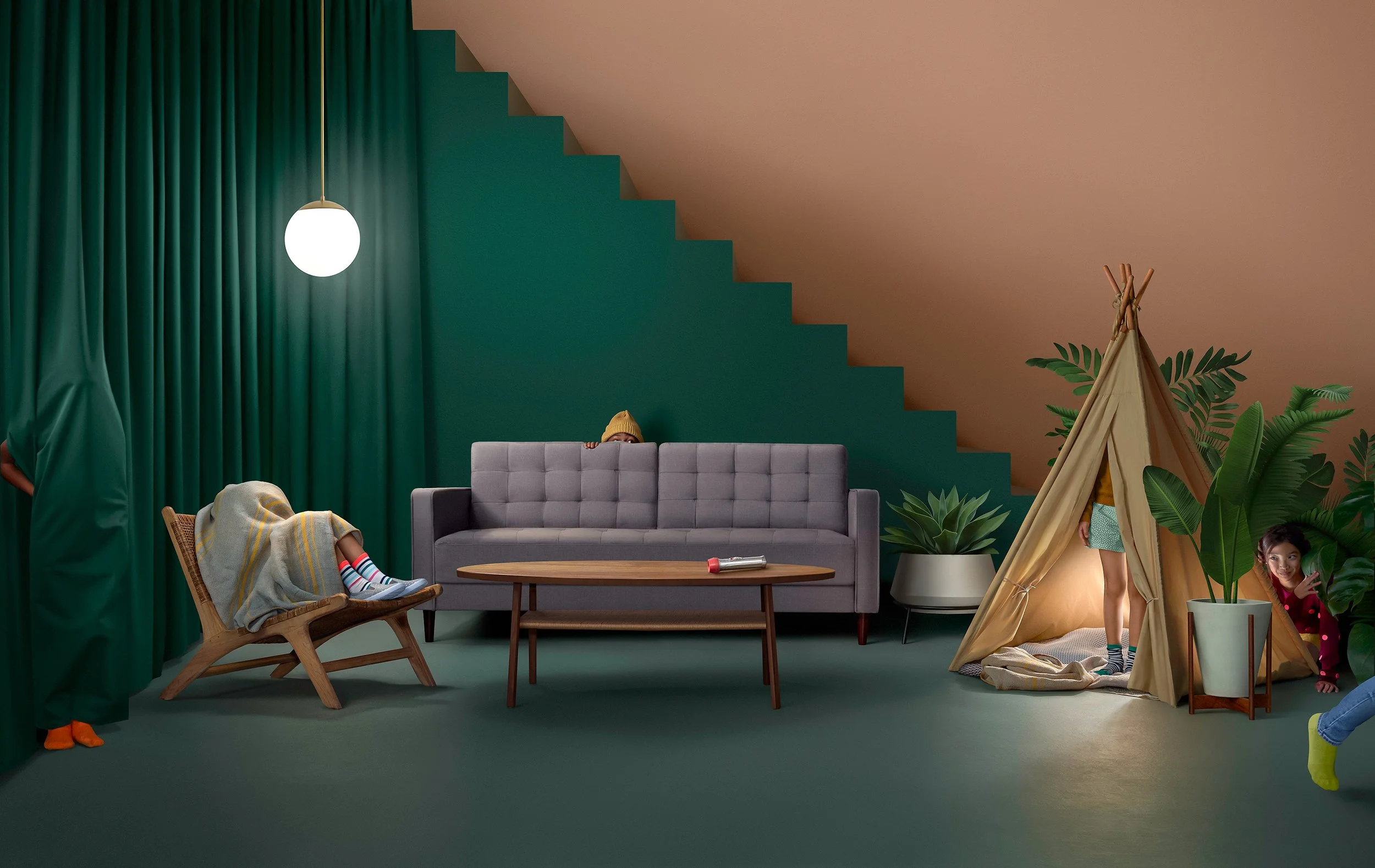 Children playing in a room with a green curtain, a gray sofa, a wooden chair with legs up, a teepee tent, and large potted plants, with a staircase in the background.