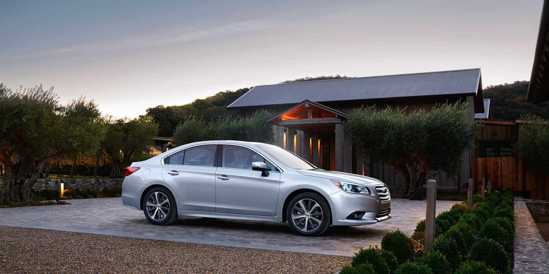 A silver sedan parked on a cobblestone driveway in front of a modern house with a wooden facade and trees around, during dusk.