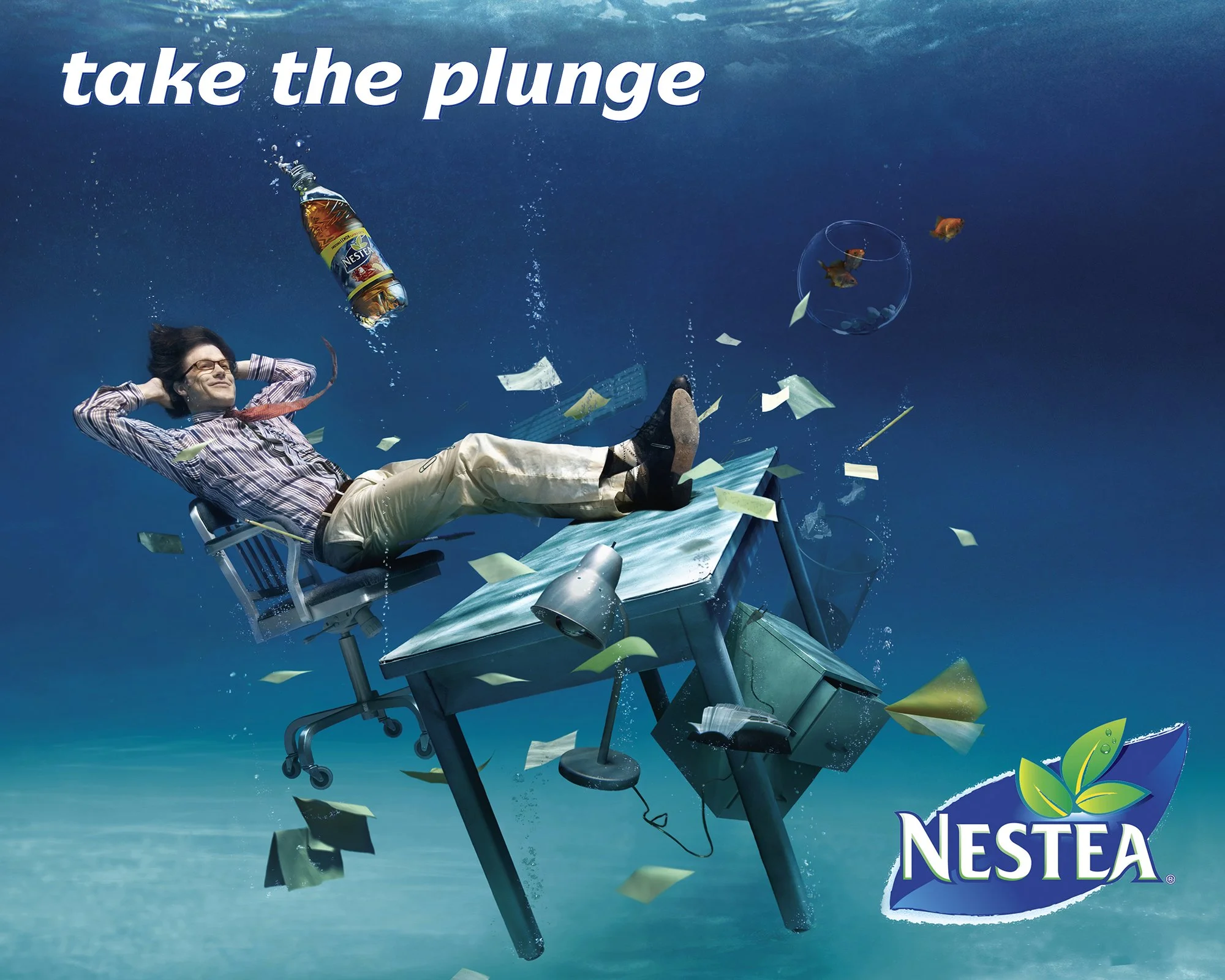 A man in office attire floating underwater at a desk, with scattered papers, a chair, a lamp, a computer monitor, a bottle of Nestea iced tea, and fish swimming around. Text on top says 'take the plunge' and Nestea logo is at the bottom right.